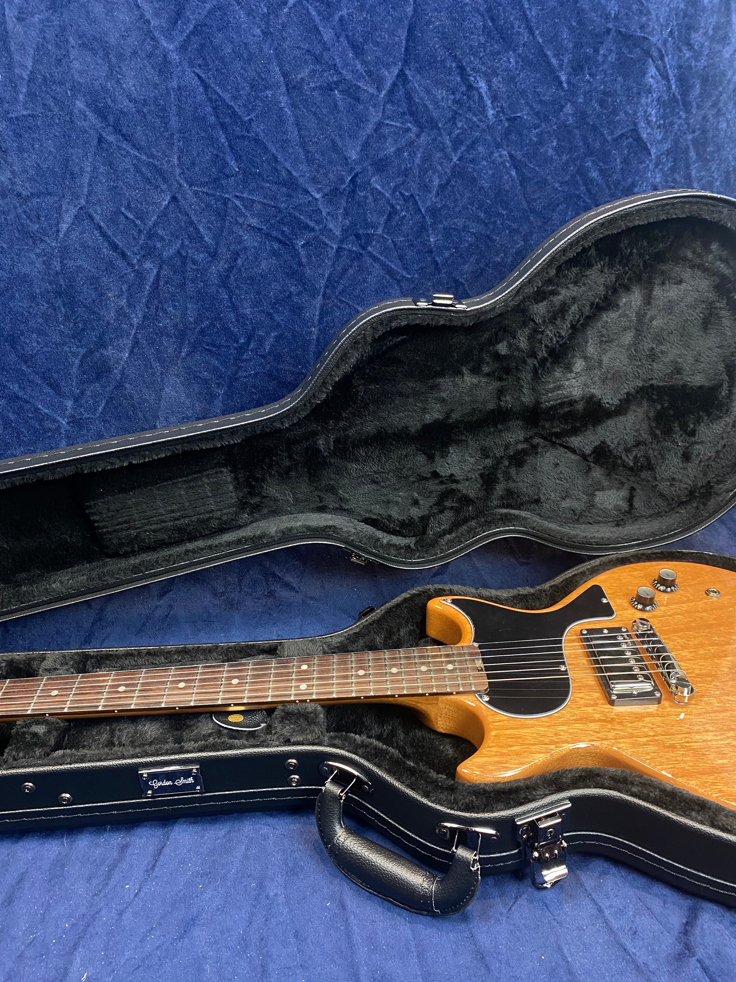 Brown electric guitar with black pickguard and chrome hardware resting in a black fur-lined hard case, case is open on a blue fabric surface.