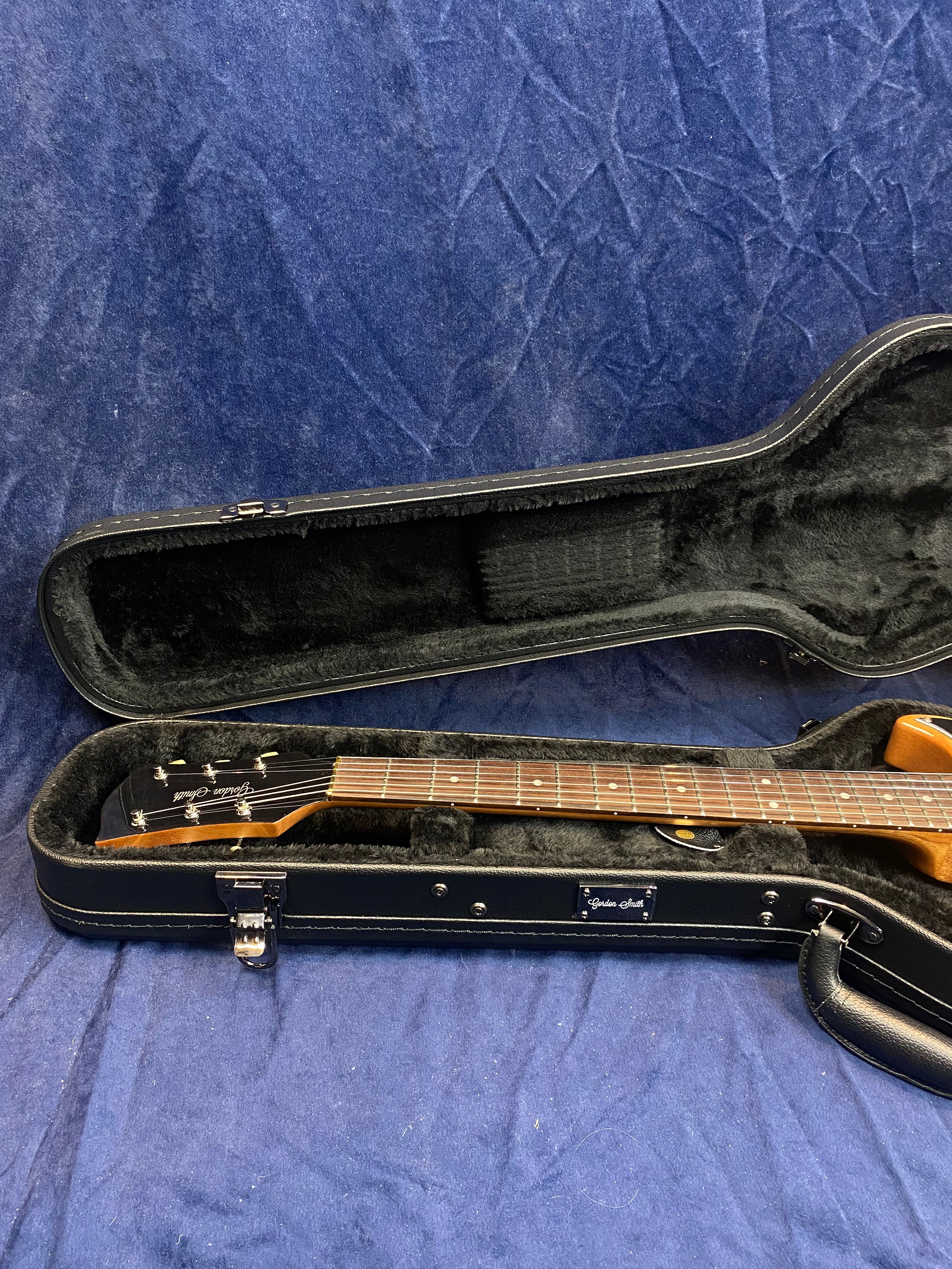 Electric guitar in a black, plush-lined hard case, lid open, showing the neck, headstock, and part of the natural wood body.