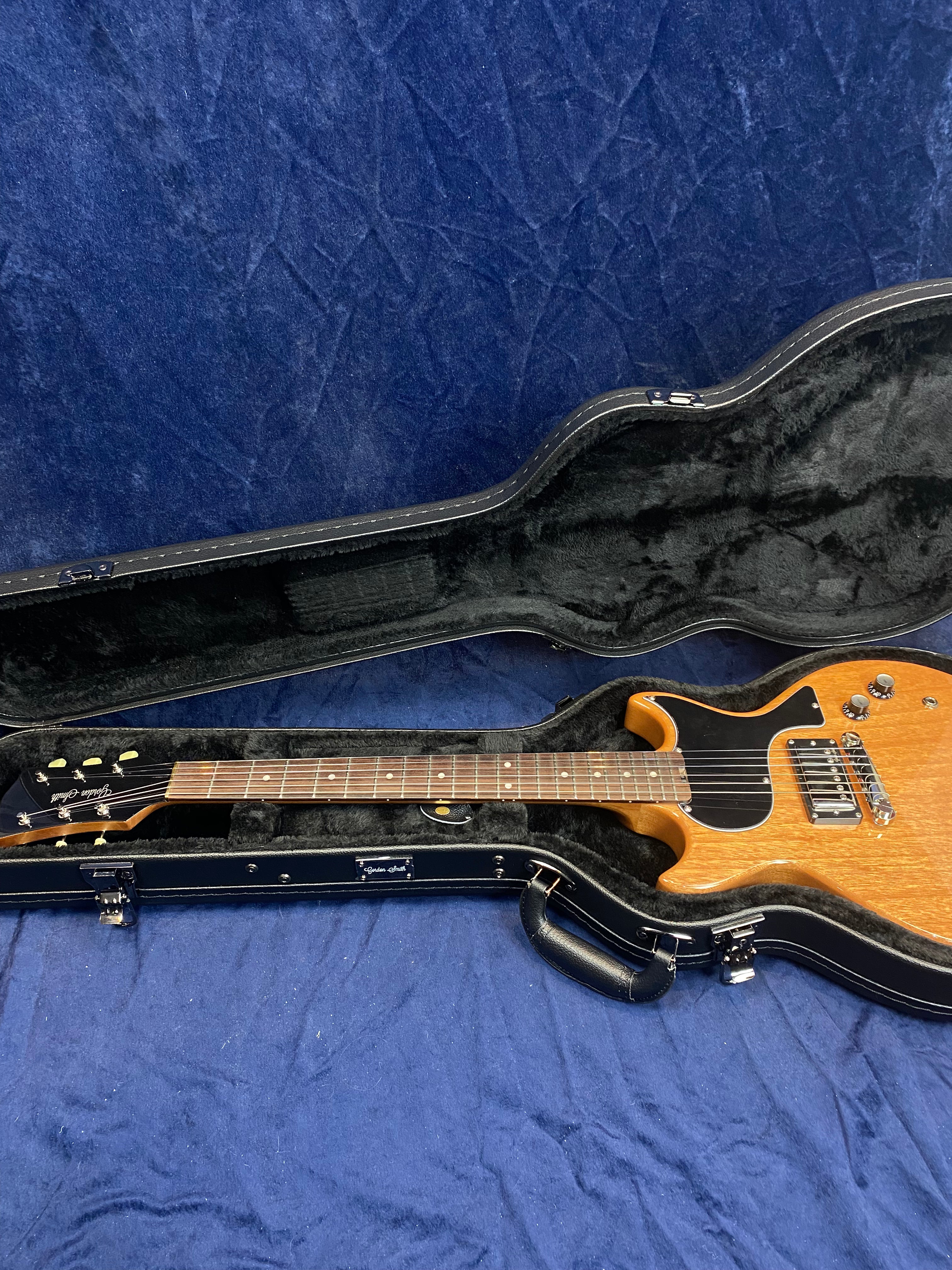 A brown solid-body electric guitar with a black pickguard rests inside a fitted black hard case lined with soft material. The case is open on blue fabric.