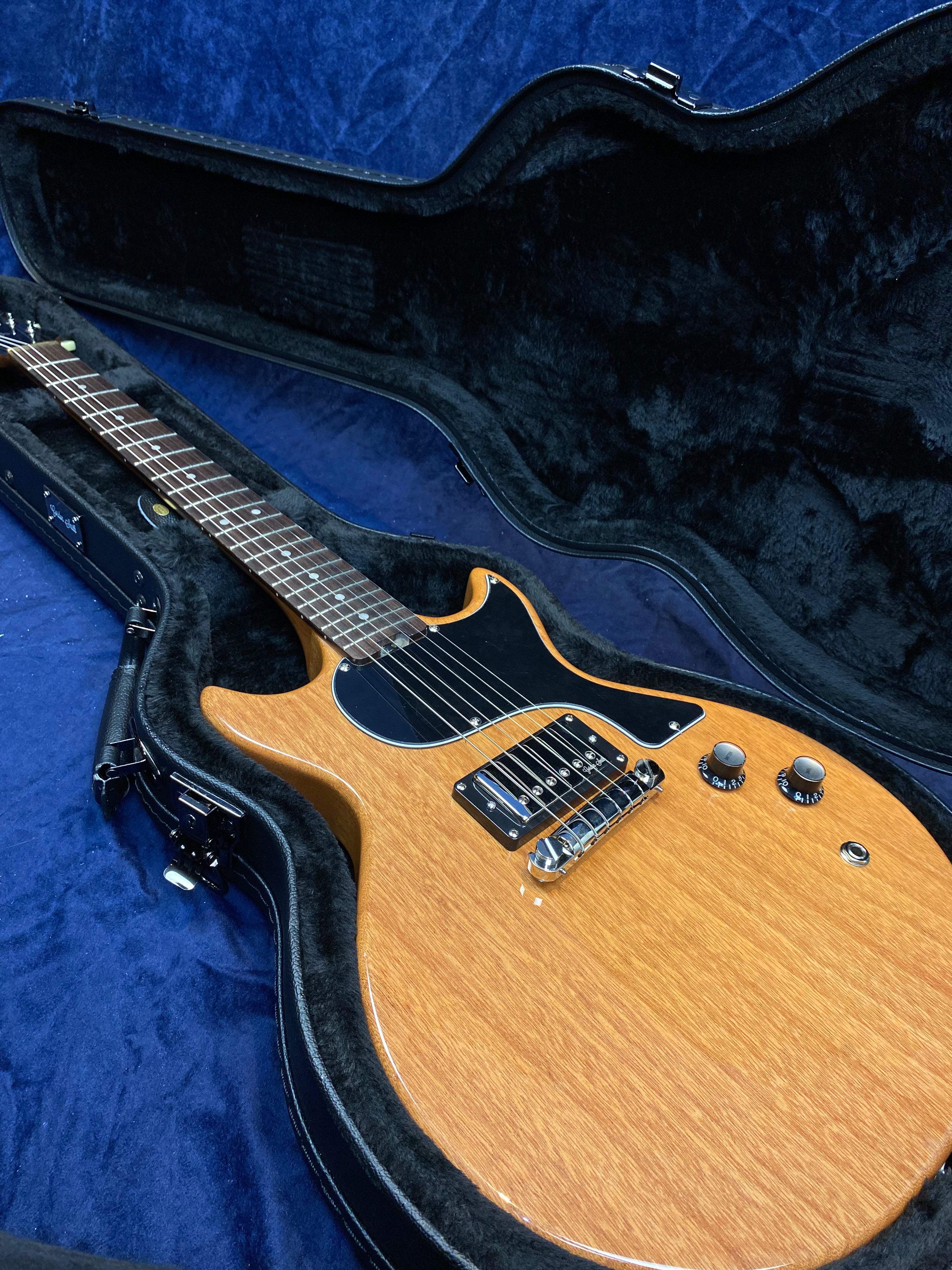 Natural wood finish electric guitar with black pickguard, two control knobs, and chrome bridge, resting inside a black padded hard case.