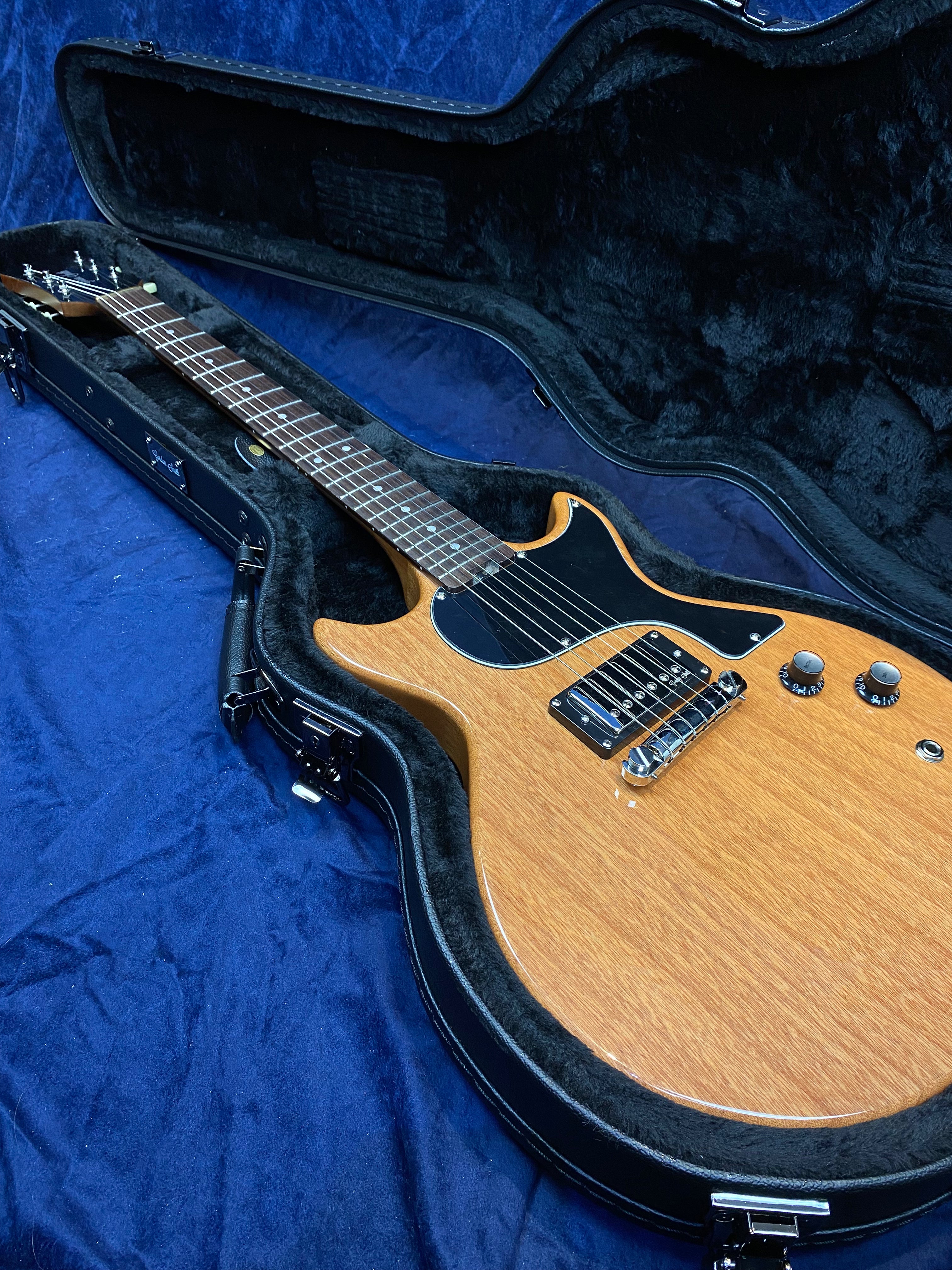 Natural wood finish electric guitar with black pickguard and two control knobs, resting inside a fitted black hard case lined with plush material.