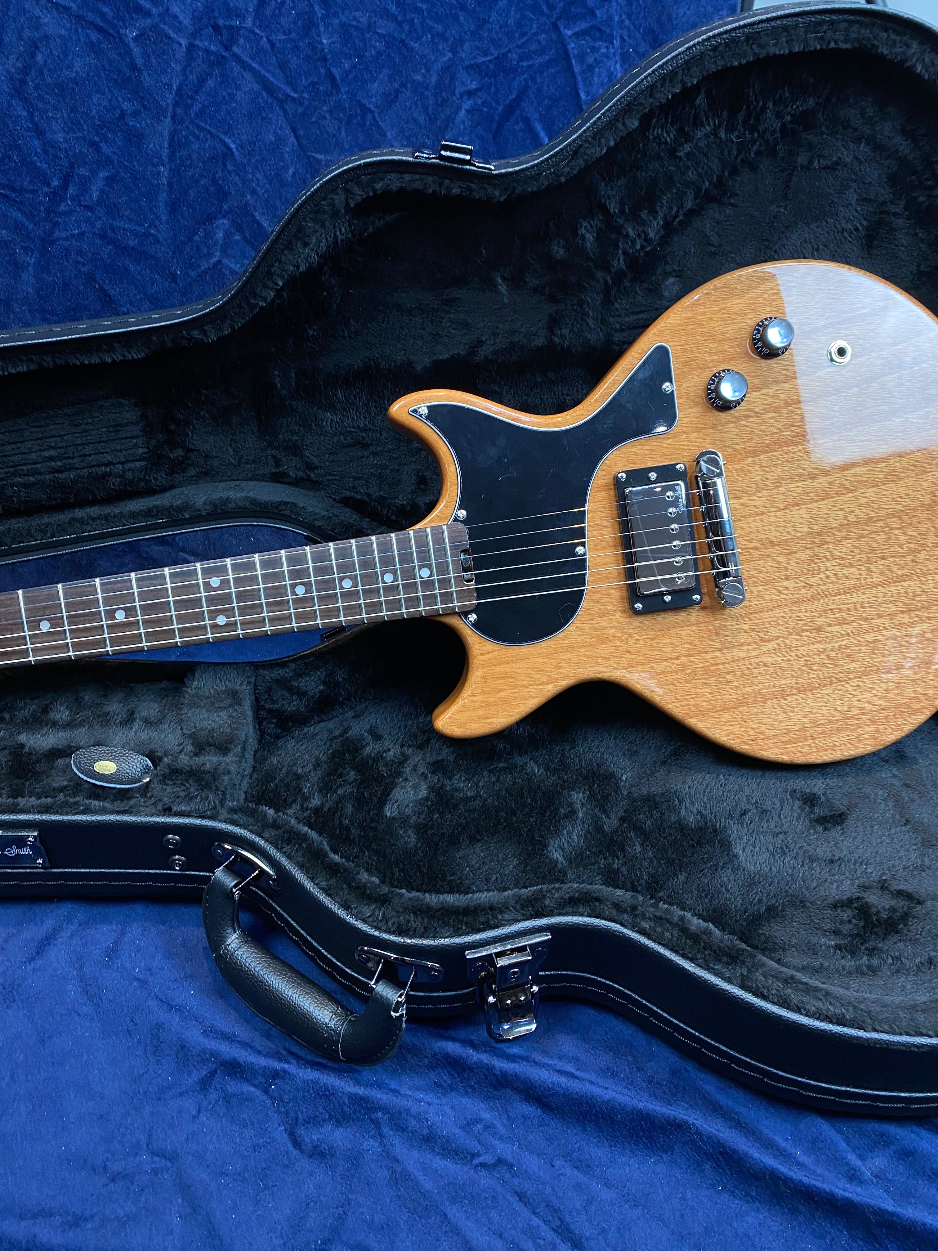 Electric guitar with a natural wood finish, black pickguard, and two control knobs resting in a black plush-lined hard case.