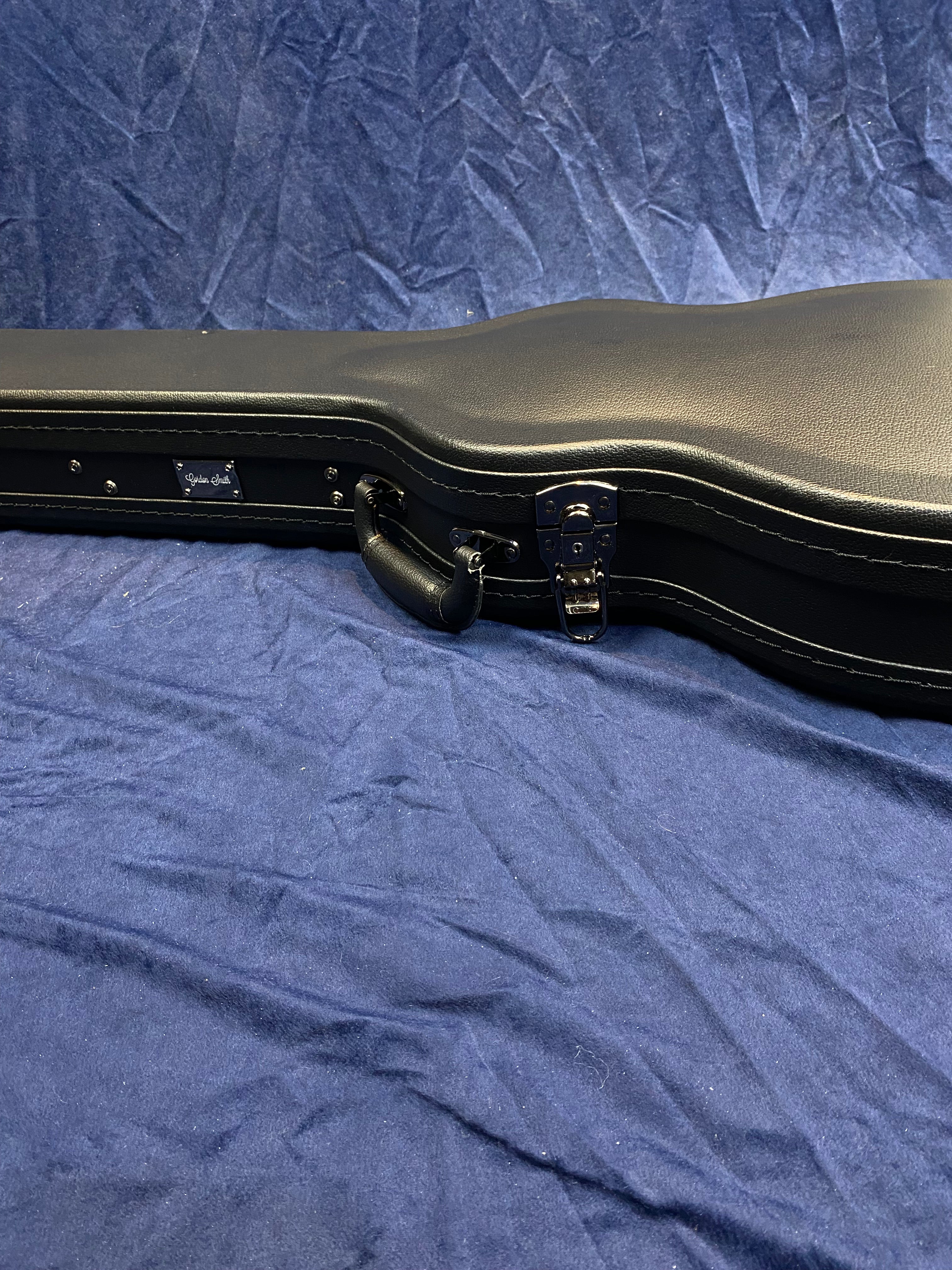Black hard case for electric guitar with metal latches and a handle, resting on a dark blue fabric background.