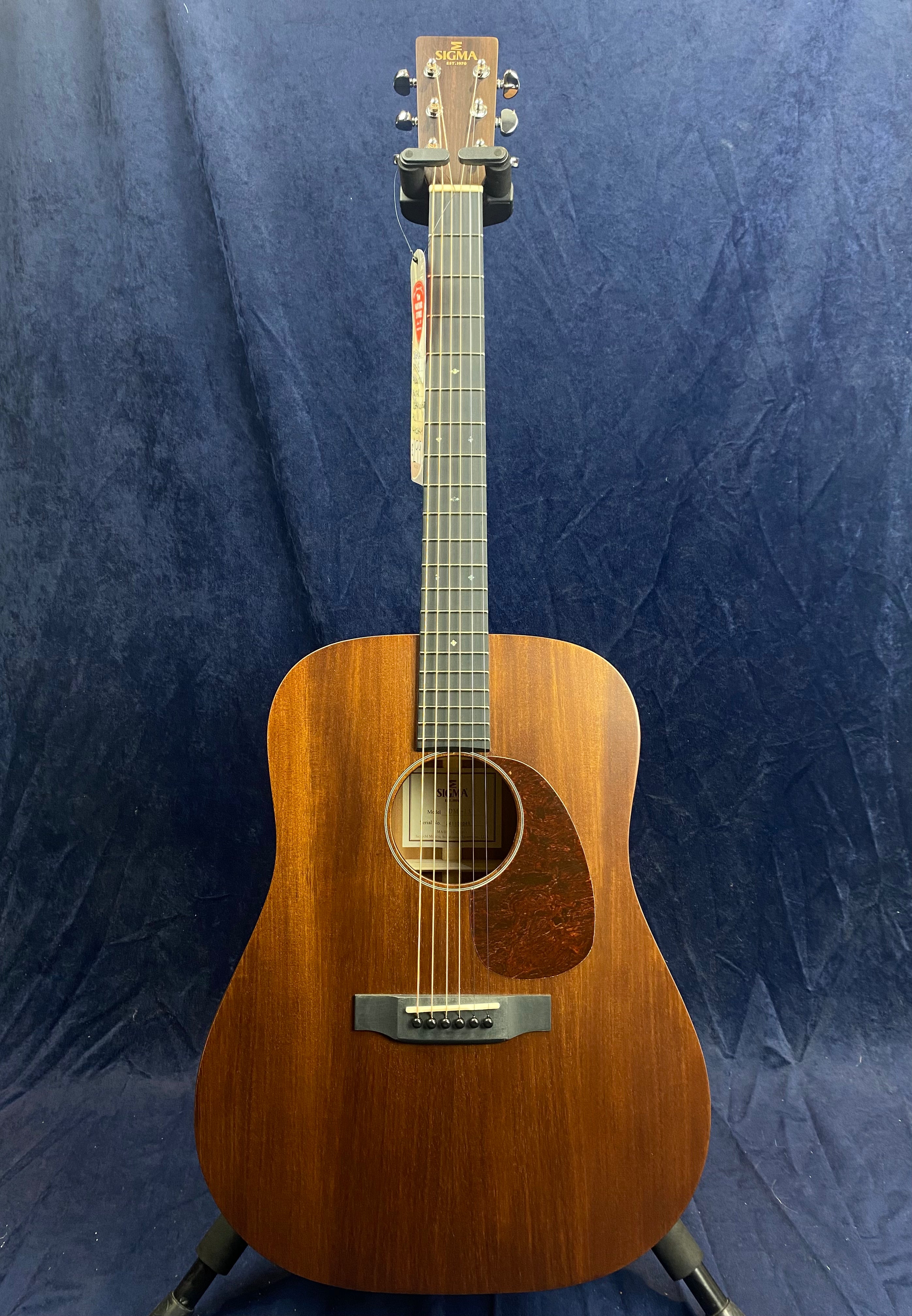 Sigma acoustic guitar with a dark wood finish, six strings, a tortoiseshell pickguard, and simple dot inlays on the fretboard, shown on a stand.