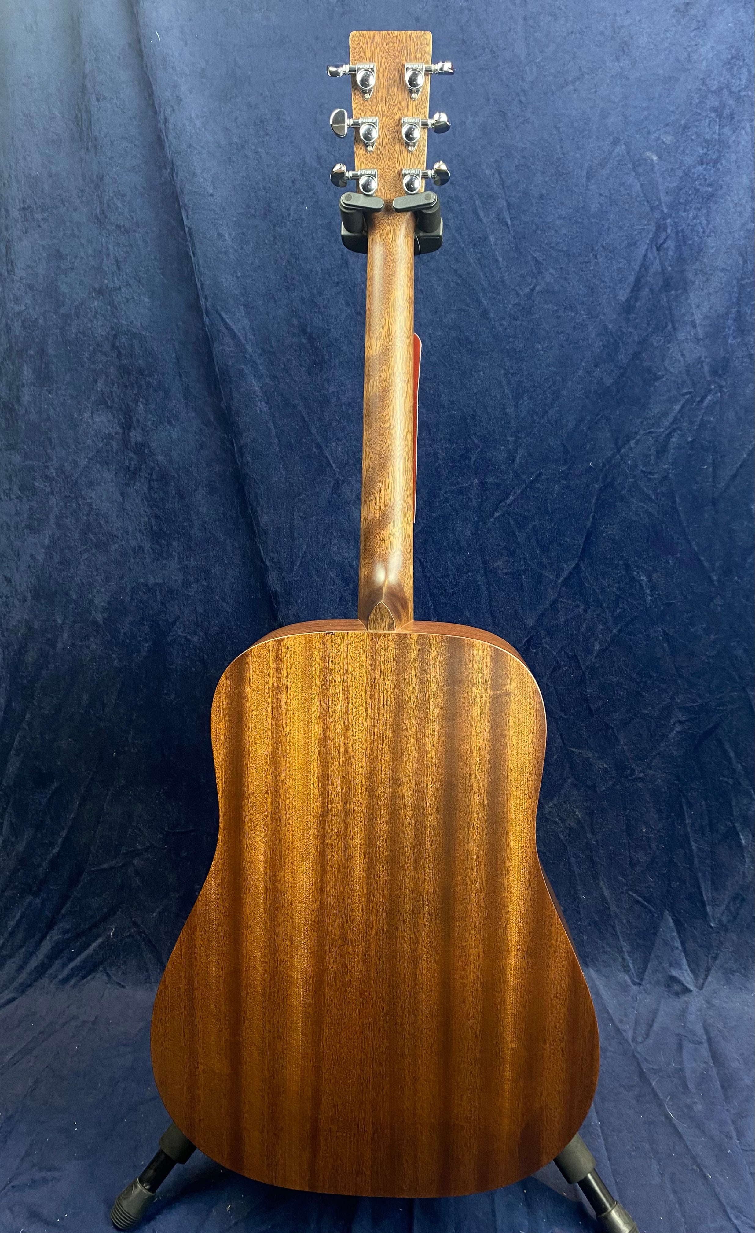 Rear view of an acoustic guitar with a smooth, dark brown wood finish, visible tuning pegs, and the instrument resting on a stand against a dark backdrop.