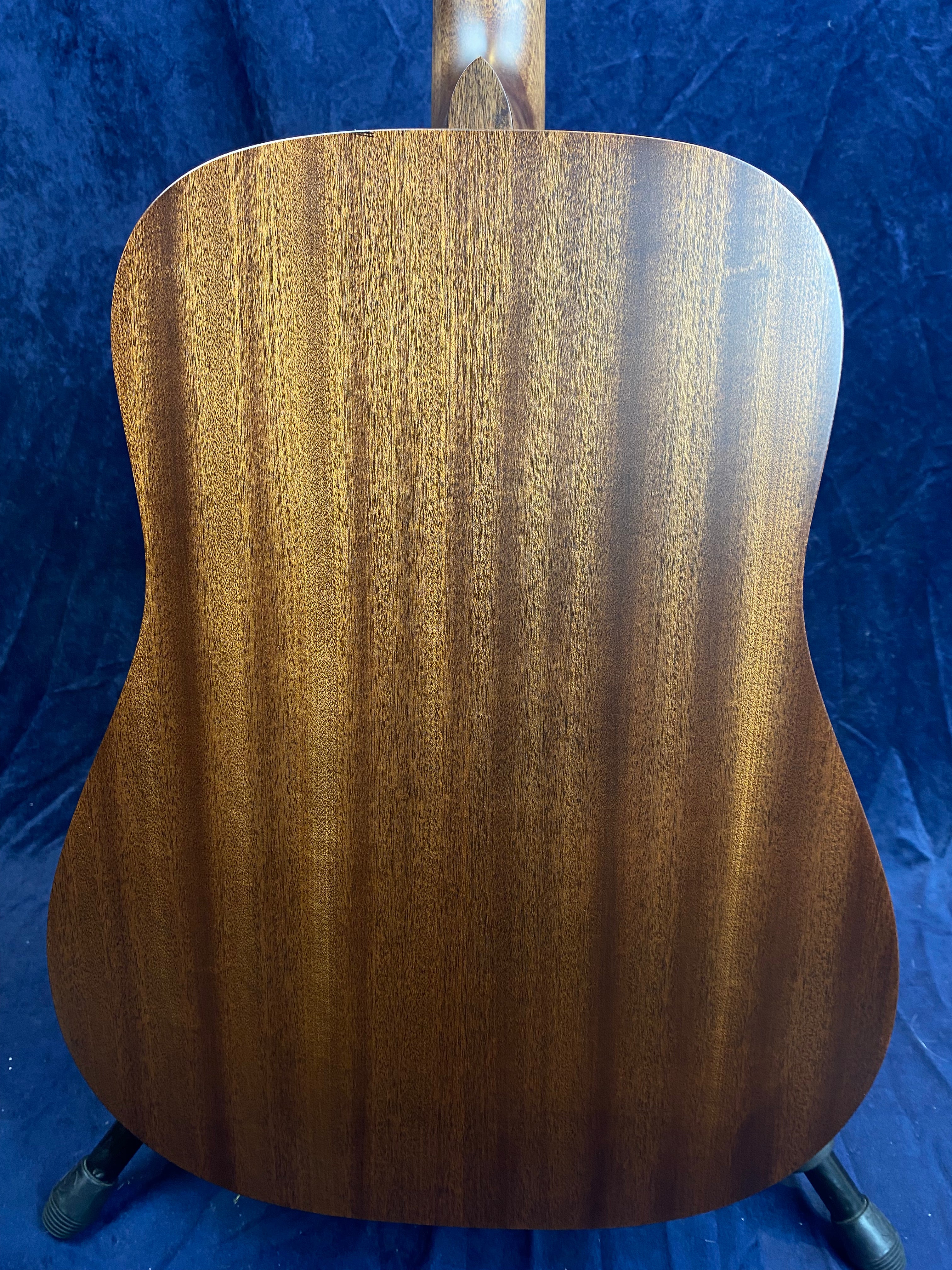 Back view of an acoustic guitar showing smooth, rich brown wood with subtle vertical grain patterns against a dark blue background.
