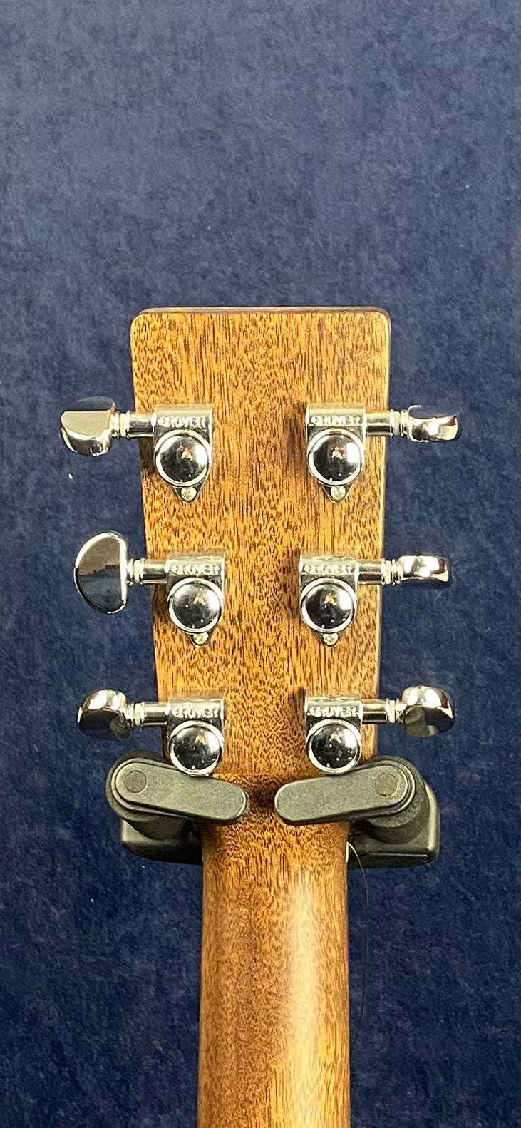 Six chrome Grover tuning pegs on the back of a wooden acoustic guitar headstock, set against a dark blue background.