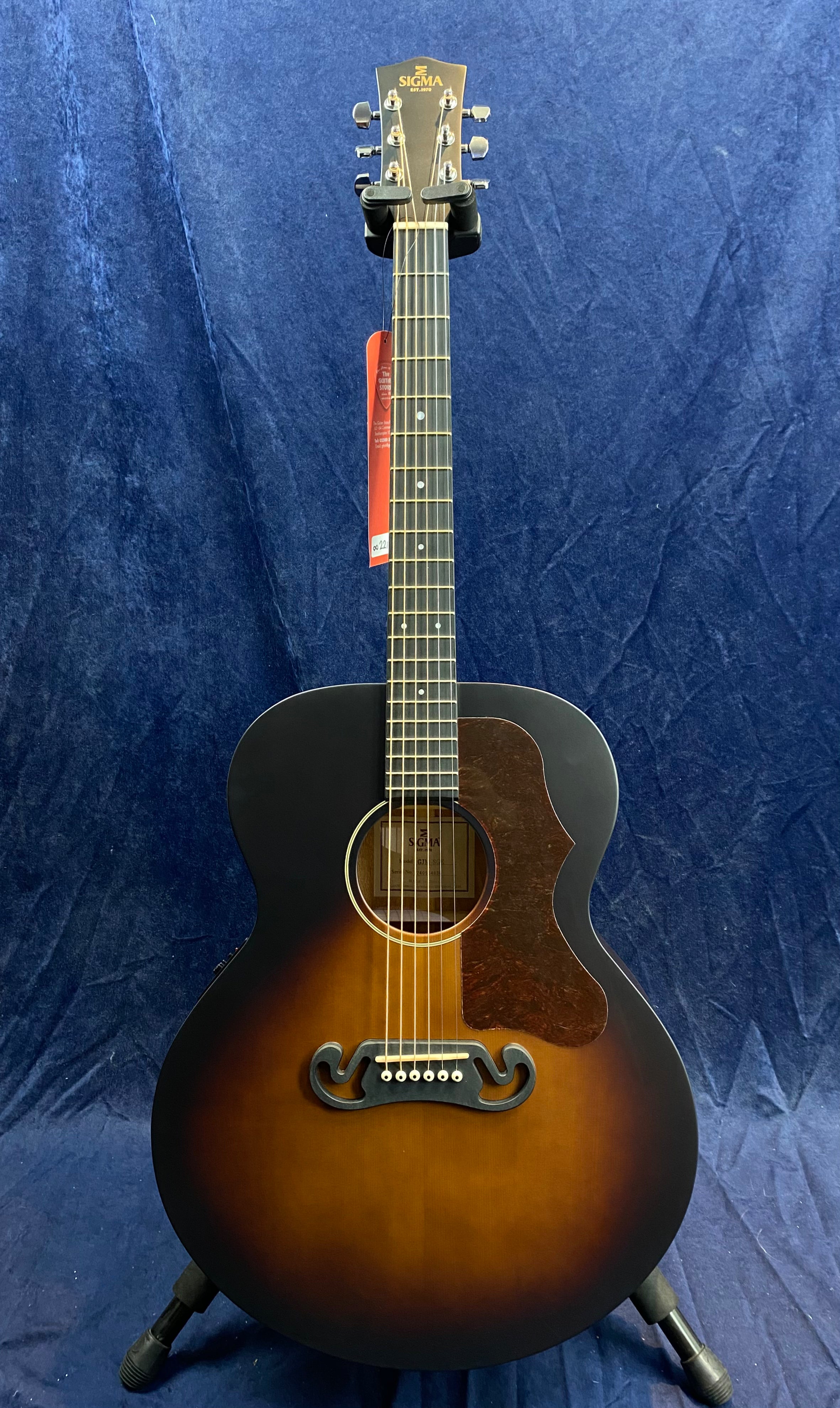 Sigma acoustic guitar with a sunburst finish, pickguard, six strings, and a decorative black bridge, displayed upright on a guitar stand.