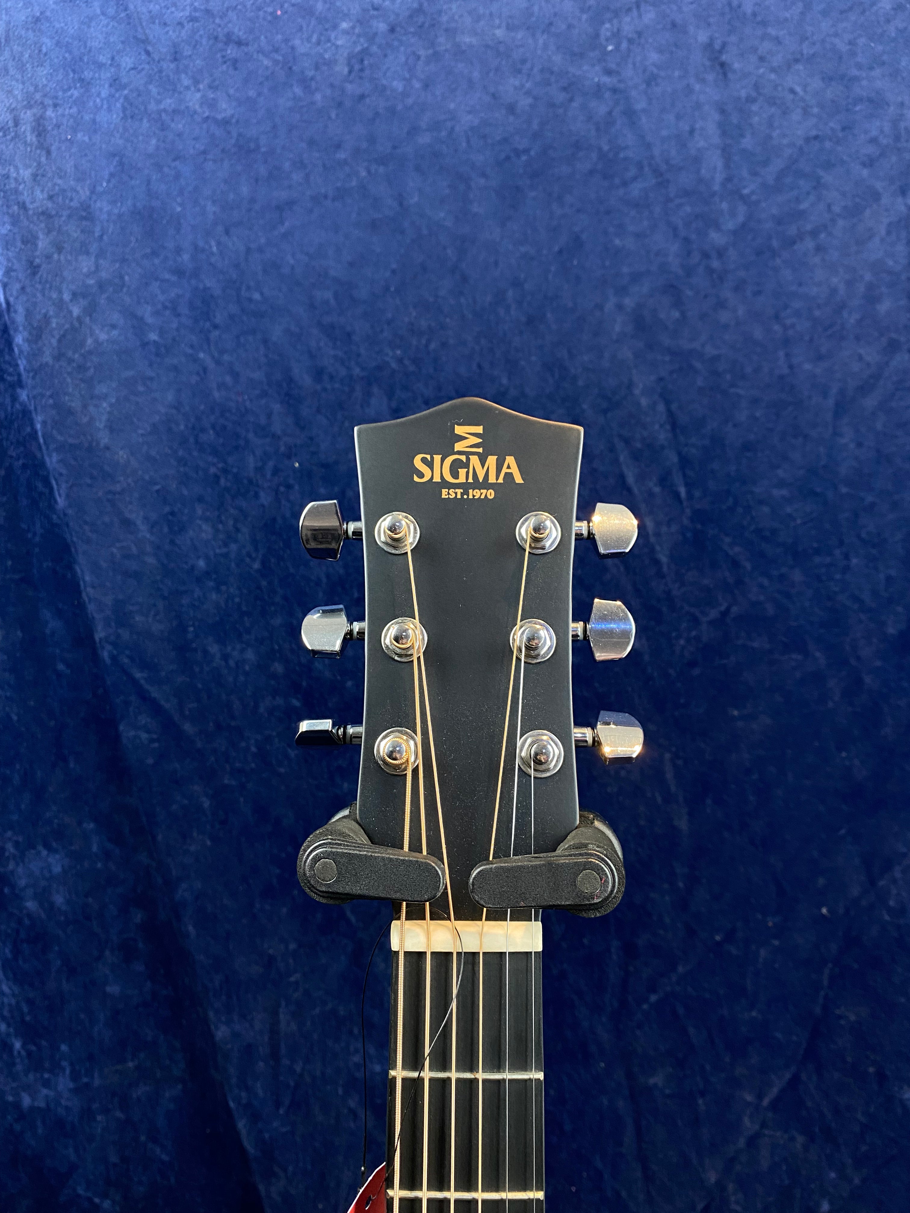 The headstock of a Sigma acoustic guitar with six tuning pegs, gold "Sigma" logo, and strings attached, set against a dark blue textured background.