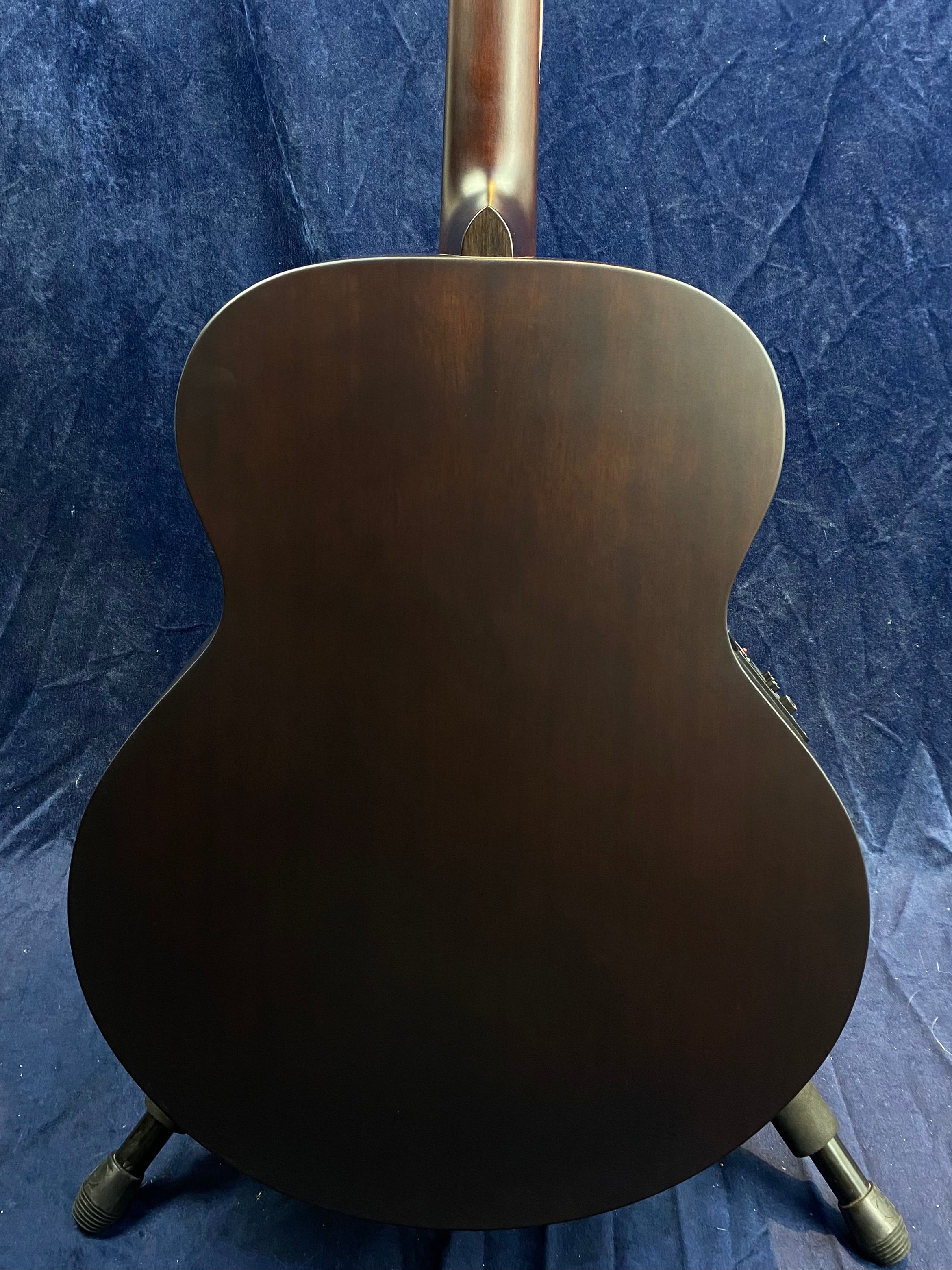 Back view of a dark brown acoustic guitar with a matte finish, resting on a stand against a dark blue textured background.