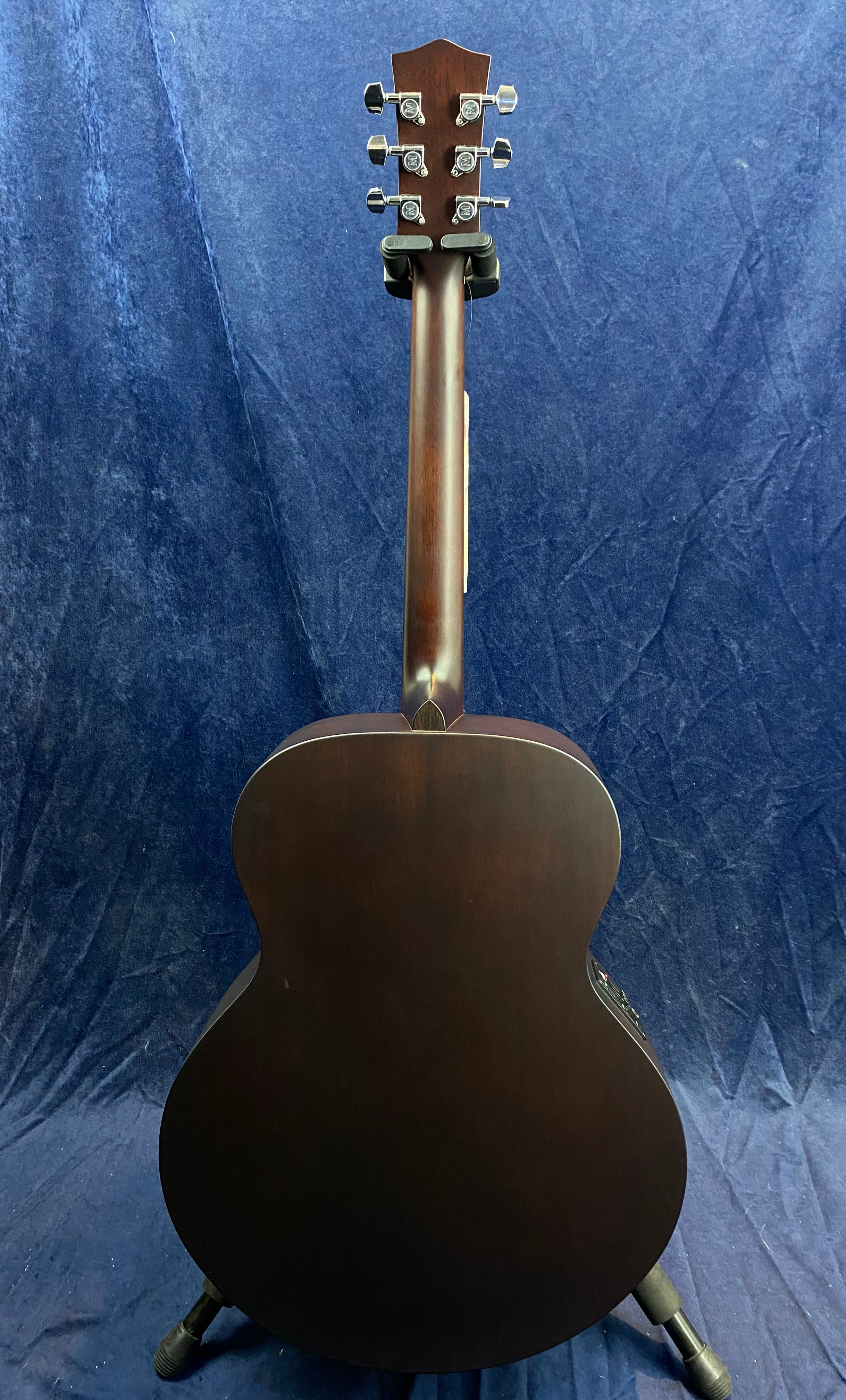 A dark wood acoustic guitar shown from the back, with six metal tuning pegs and a matte finish, resting upright on a stand against a blue backdrop.