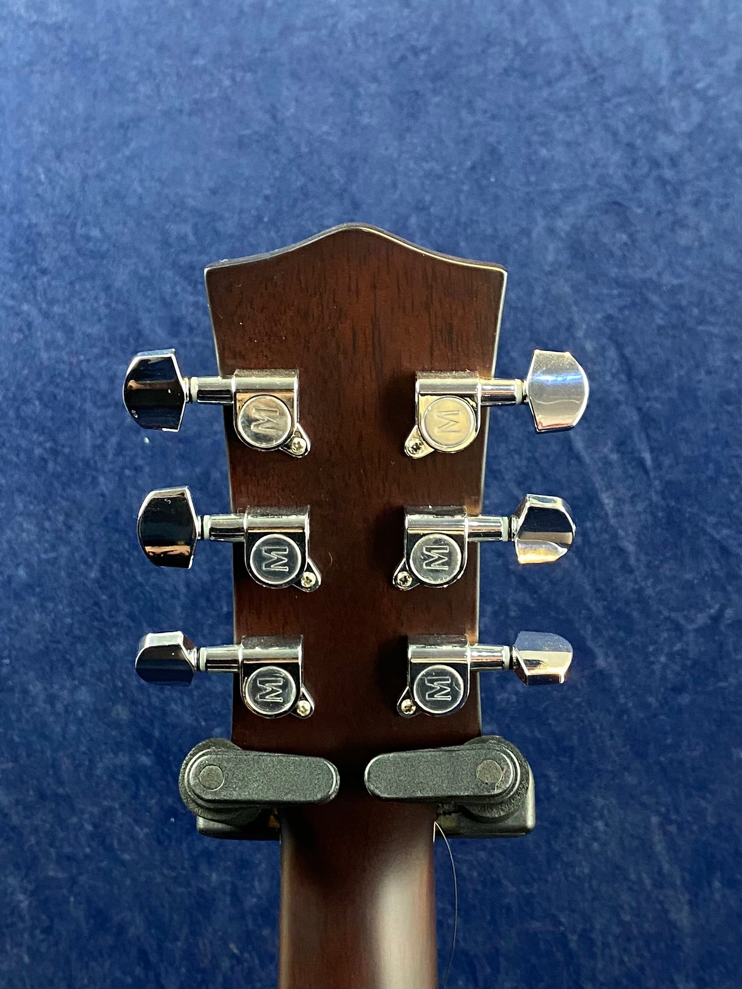Six chrome tuning pegs with "W" logos are mounted on the back of a dark wooden acoustic guitar headstock against a blue textured background.