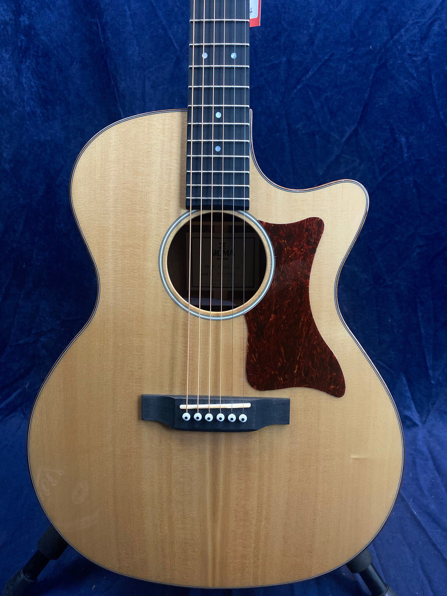 Light wood acoustic guitar with a single cutaway, six strings, a dark brown pickguard, and white bridge pins against a dark blue background.