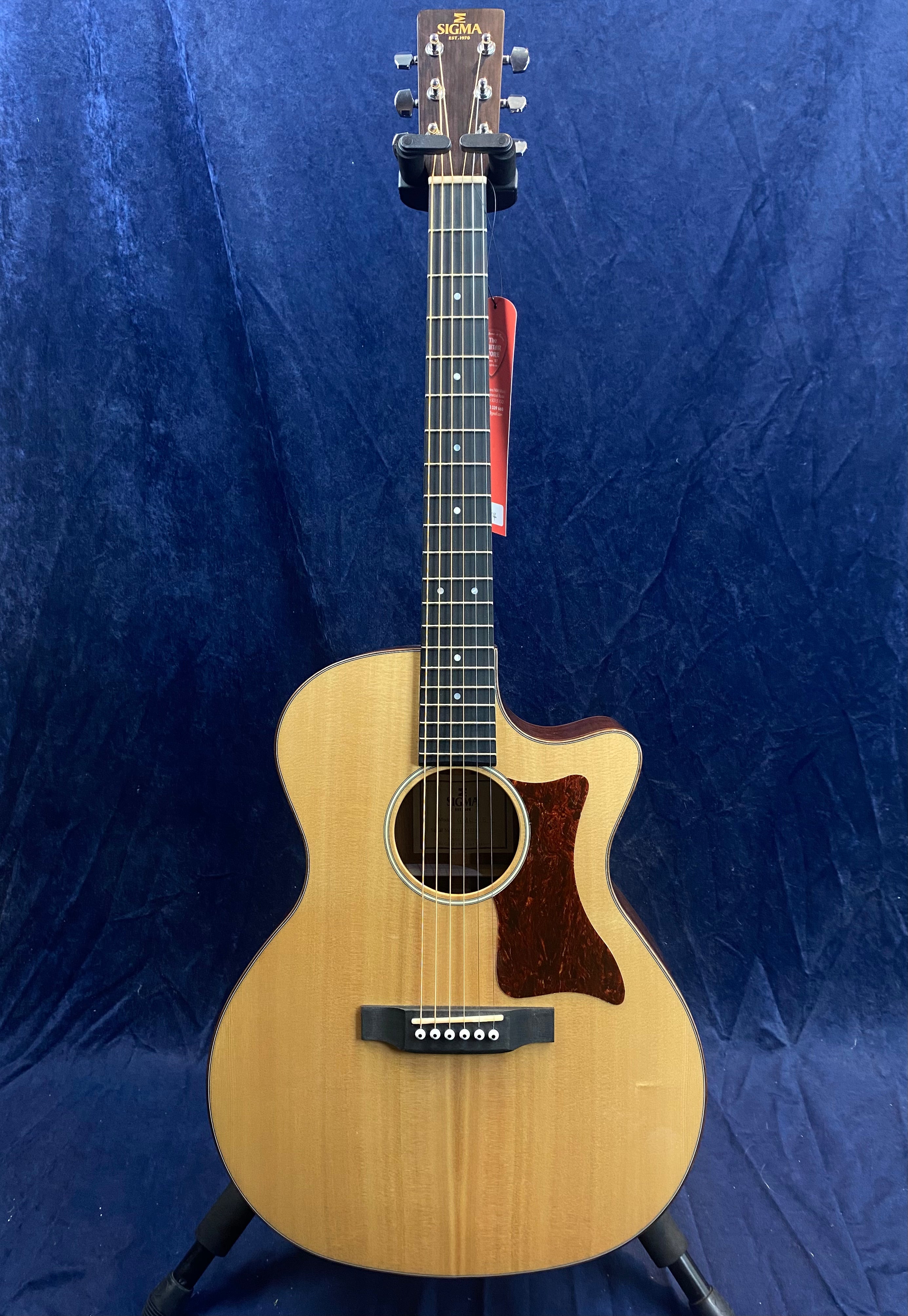 Natural wood acoustic guitar with a cutaway body, dark pickguard, six strings, and "Sigma" brand on the headstock, shown against a blue background.