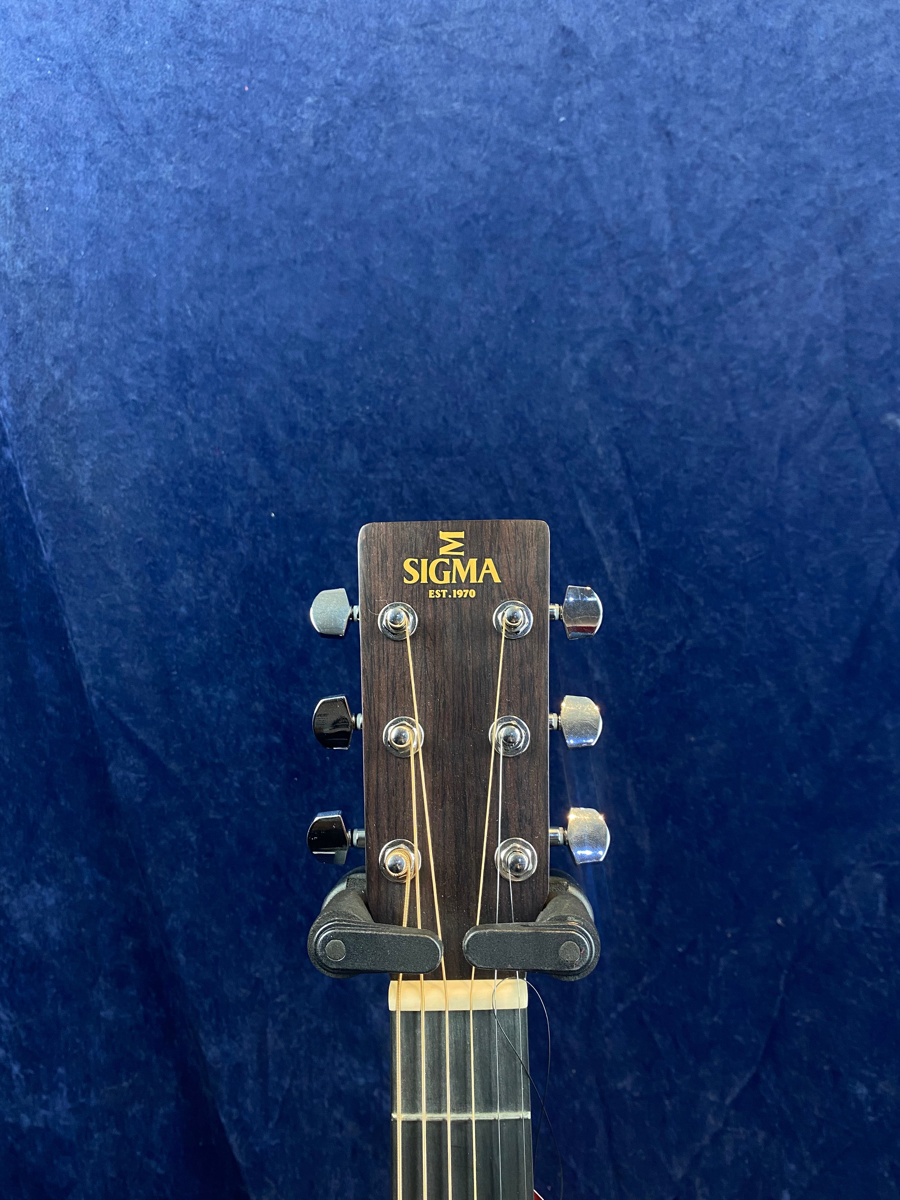 Headstock of a Sigma acoustic guitar with six tuning pegs, brown wood finish, and “SIGMA EST. 1970” in gold lettering, against a dark blue background.