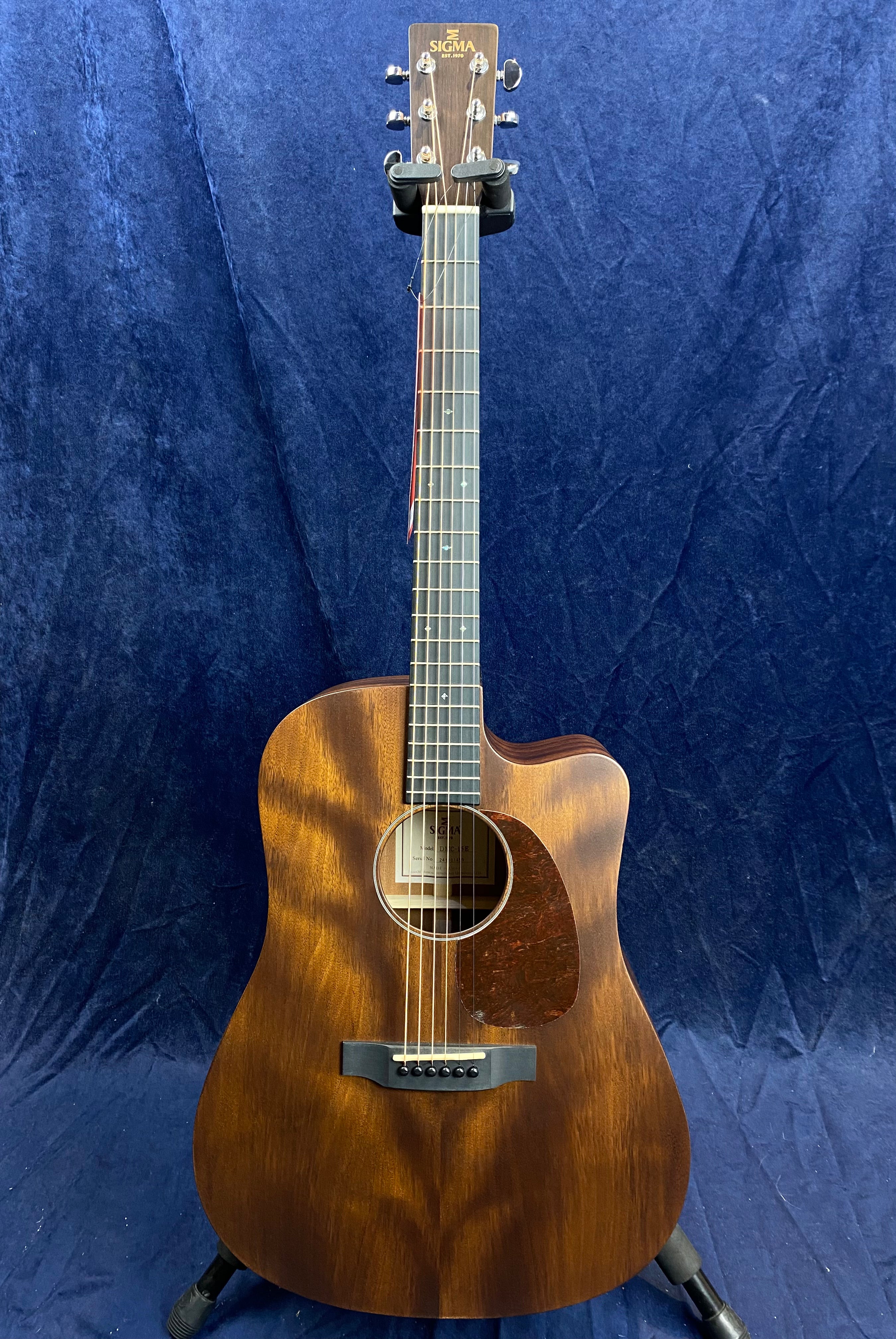 Sigma acoustic guitar with a dark wood finish, cutaway body on the right, six strings, round sound hole, and silver tuning pegs, standing on a stand.