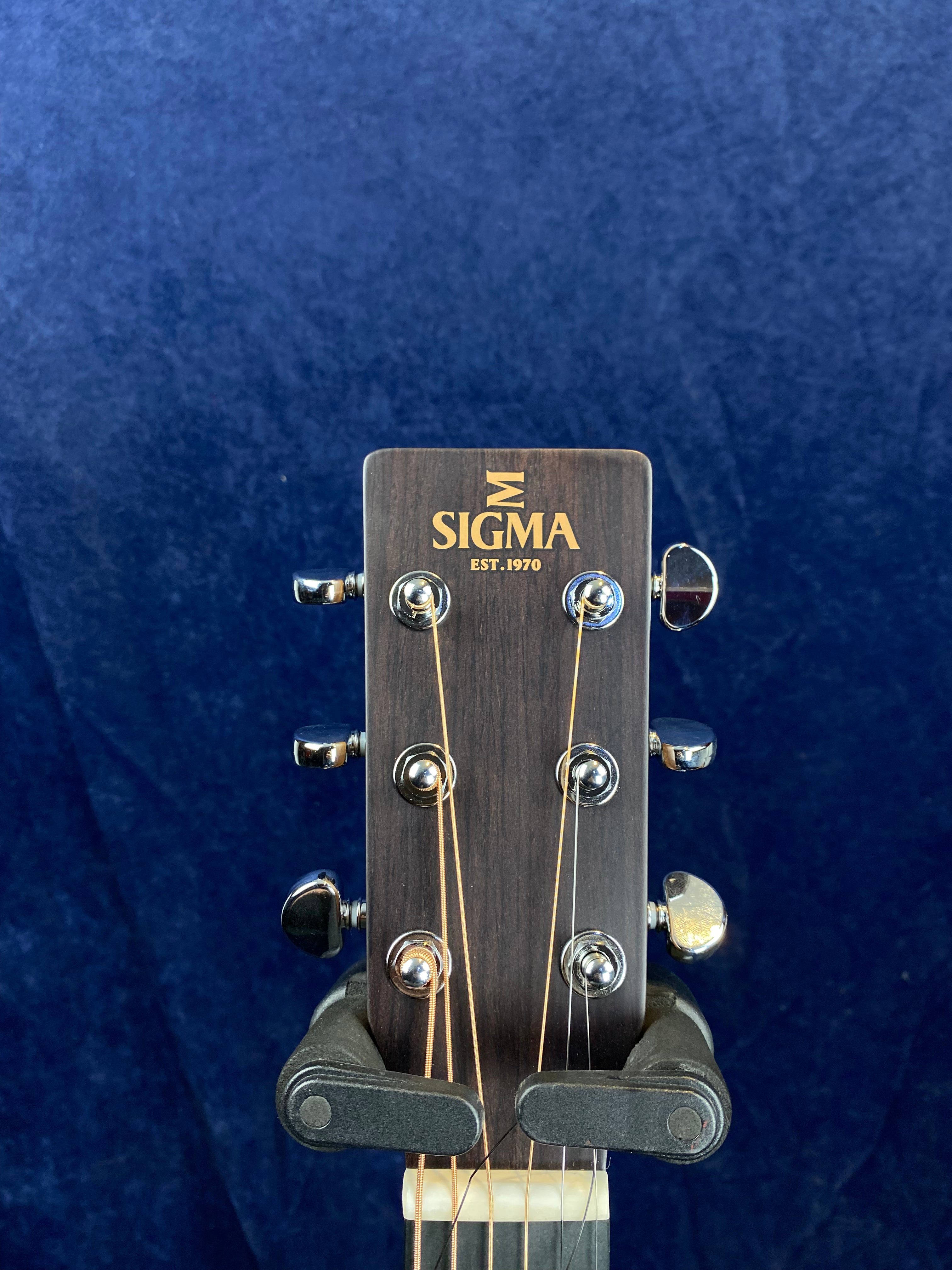 Headstock of a Sigma acoustic guitar with six tuning pegs, dark wood finish, and gold lettering reading “SIGMA EST. 1970.”