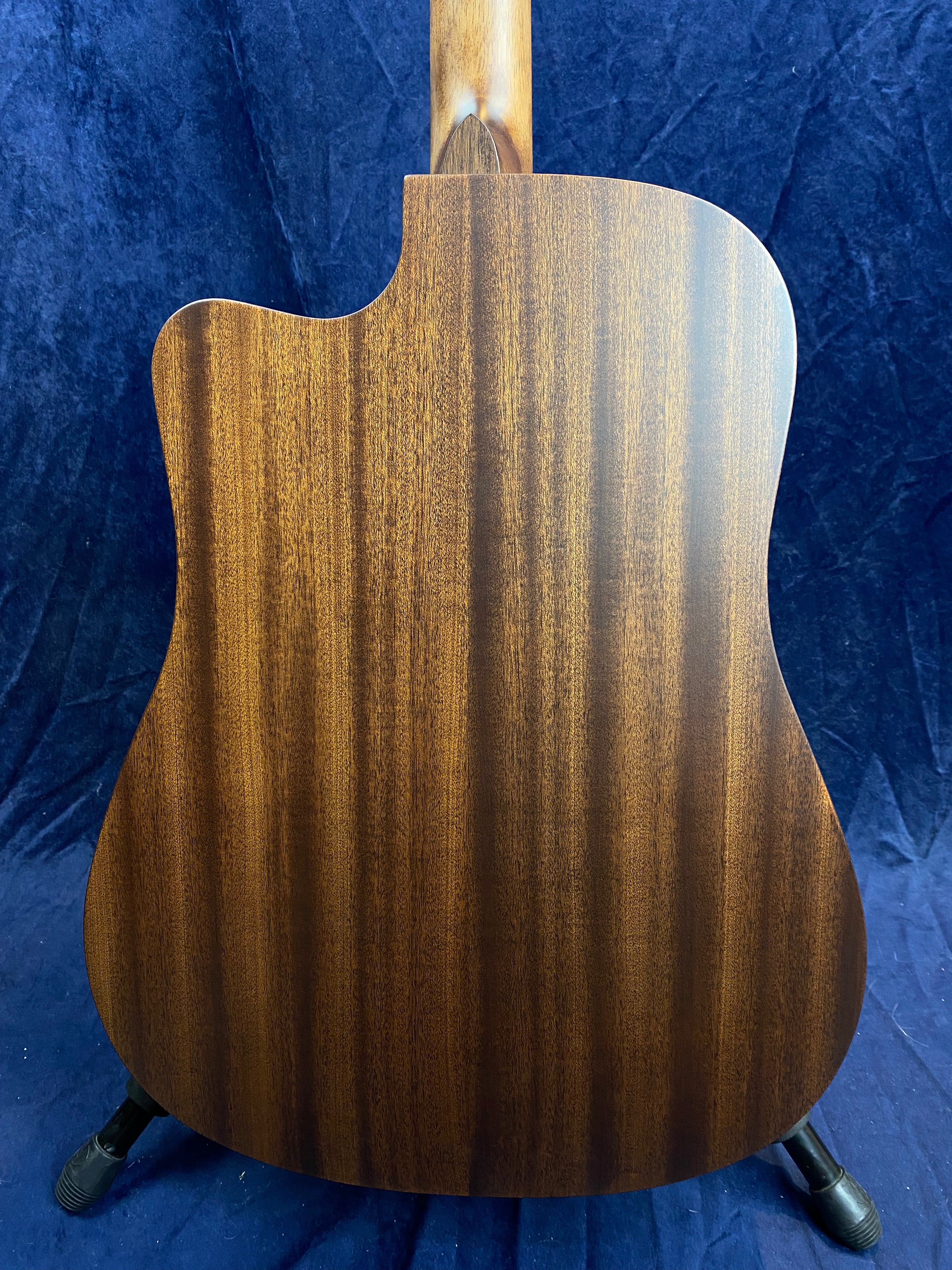 Back of an acoustic guitar with a cutaway, showing smooth, dark brown wood grain and a natural finish against a dark blue fabric background.