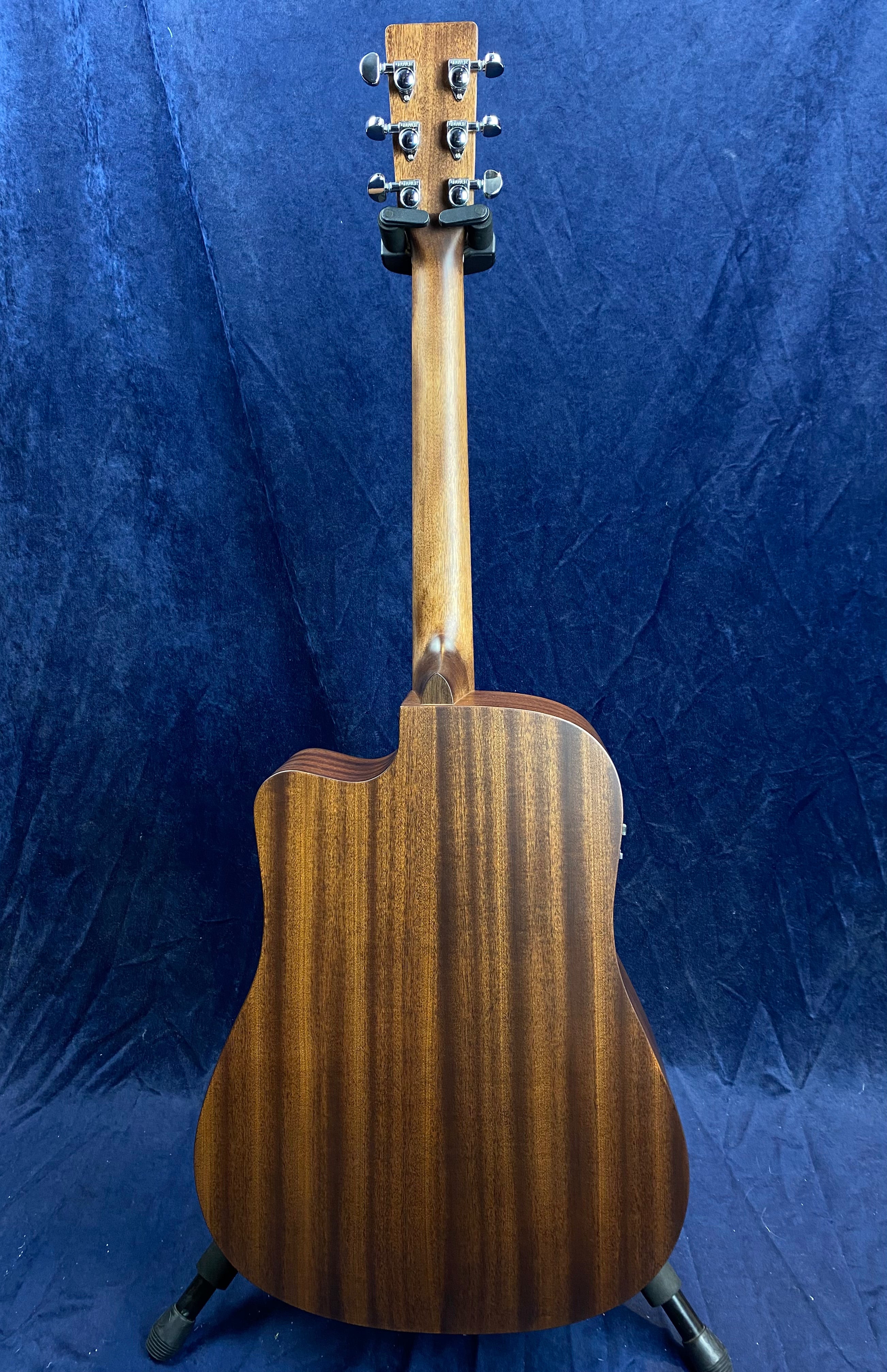 Back view of a wooden acoustic guitar with a cutaway body, vertical wood grain, and six tuning pegs, standing on a guitar stand.