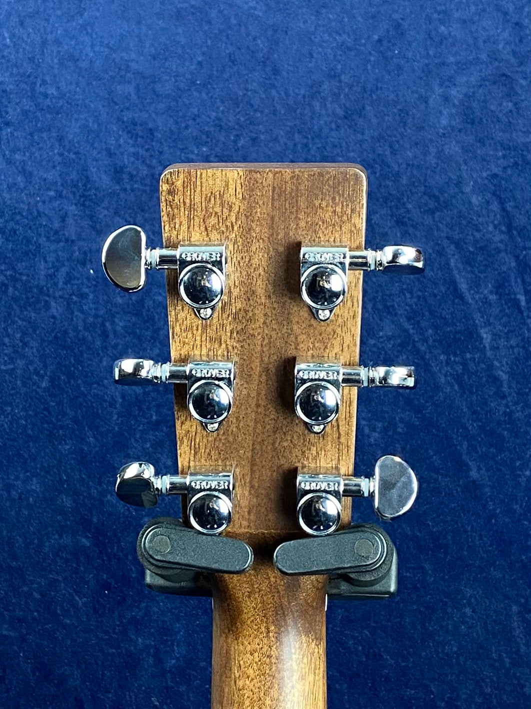 Six chrome Grover tuning pegs on the back of a wooden acoustic guitar headstock, against a dark blue background.