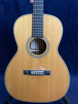 Natural wood acoustic guitar with a dark fretboard, six strings, and a simple rosette around the sound hole, shown against a dark blue background.