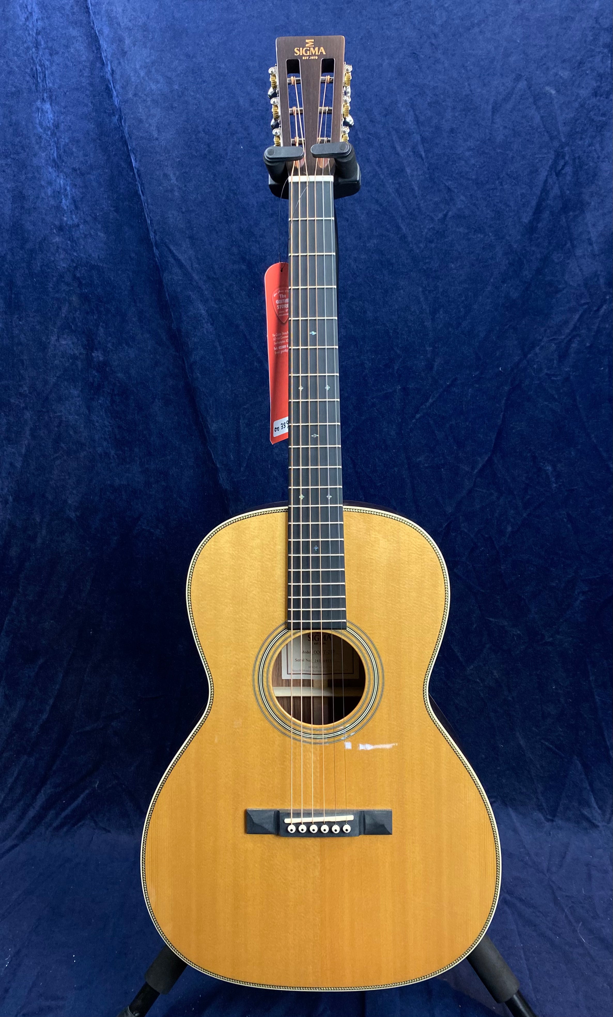 Sigma acoustic guitar with a natural wood finish, slotted headstock, and six strings, displayed upright against a dark blue background.