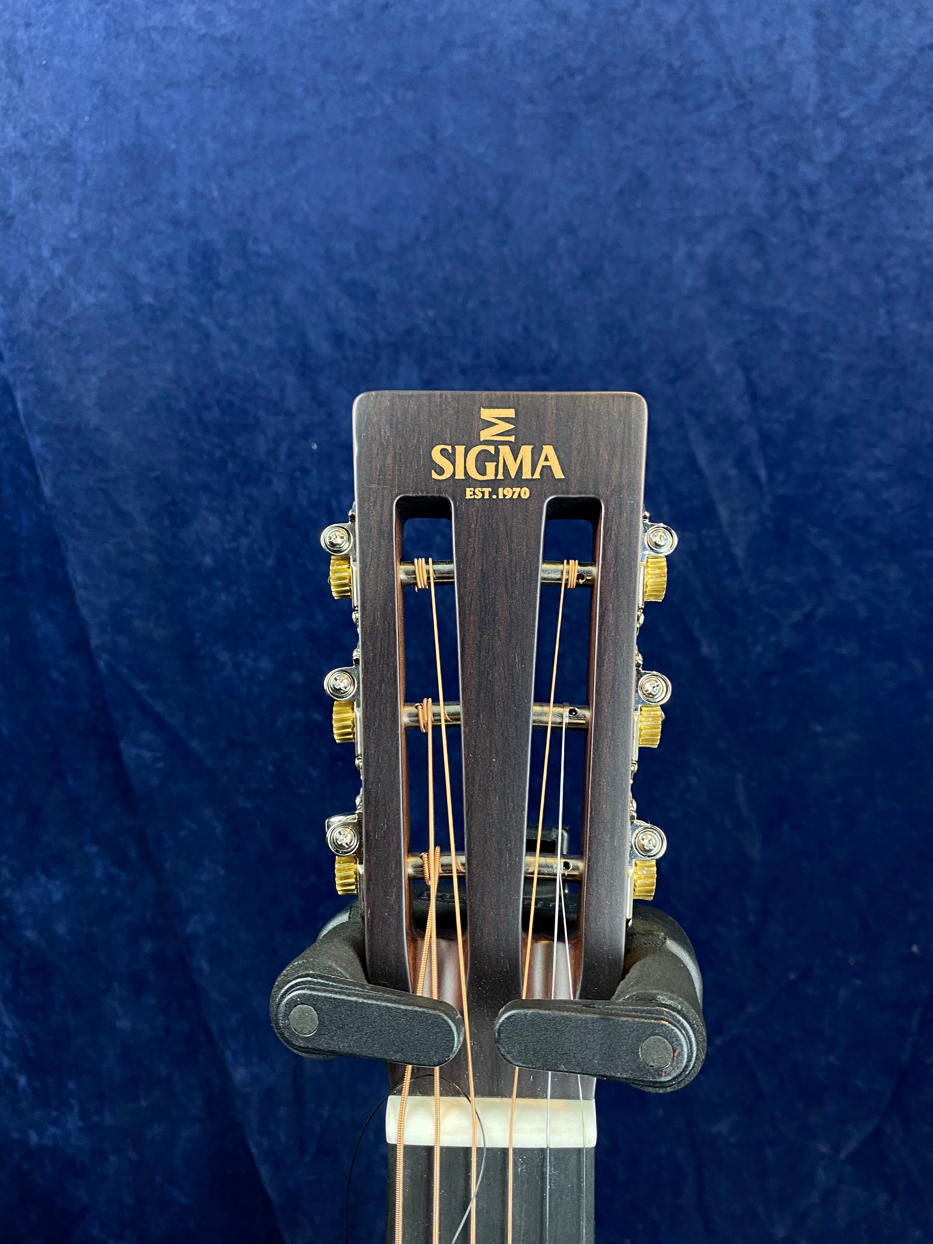 Headstock of a Sigma acoustic guitar with gold tuning pegs, brown wood finish, and the logo "SIGMA EST. 1970" in gold lettering.