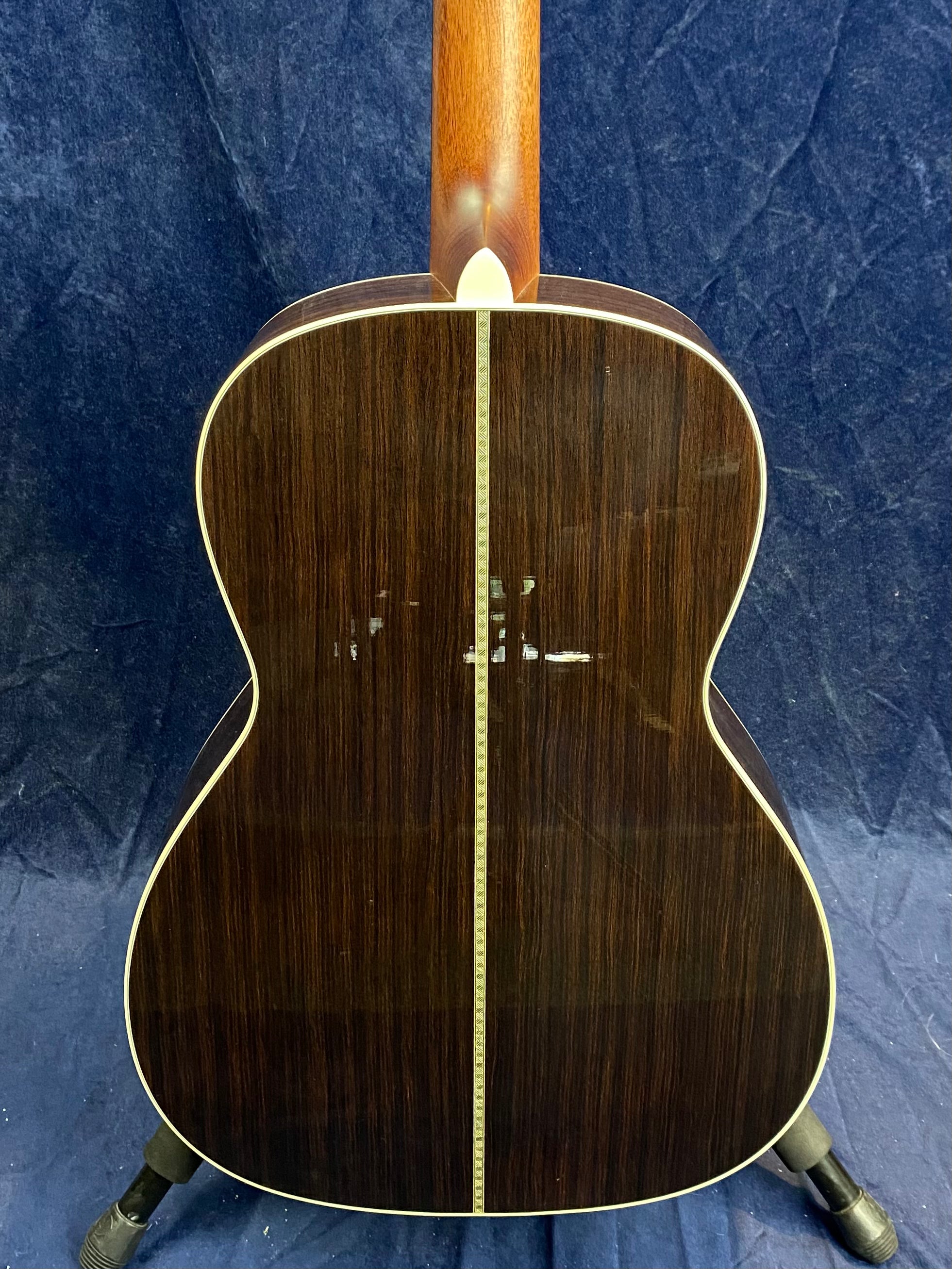 Back view of an acoustic guitar with dark wood, a light decorative center strip, cream binding, and a natural wood neck against a blue background.