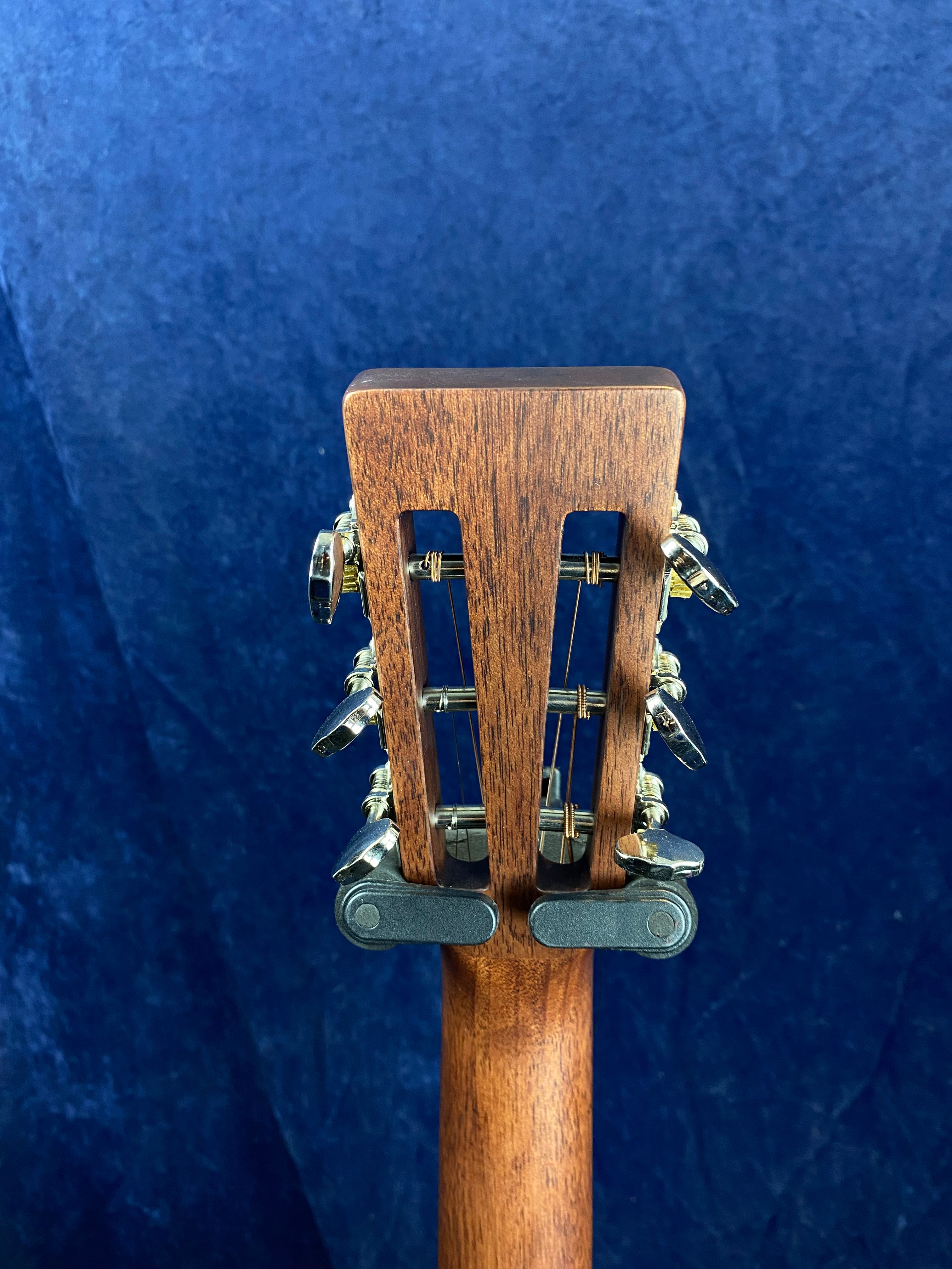 The back of a wooden acoustic guitar headstock with silver tuners and slotted openings, shown against a dark blue textured background.