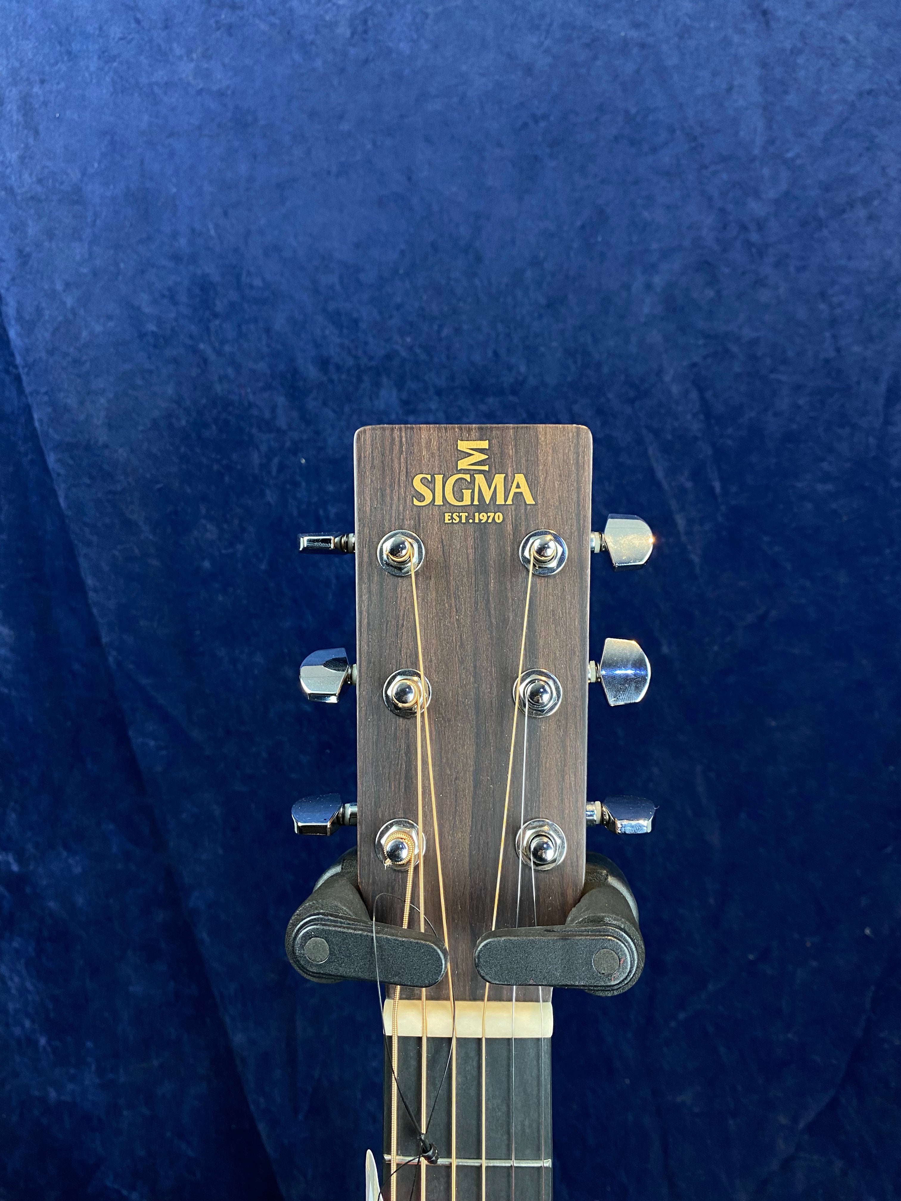 Headstock of a Sigma acoustic guitar with six tuning pegs, dark wood finish, and "SIGMA EST. 1970" in gold letters, against a blue background.
