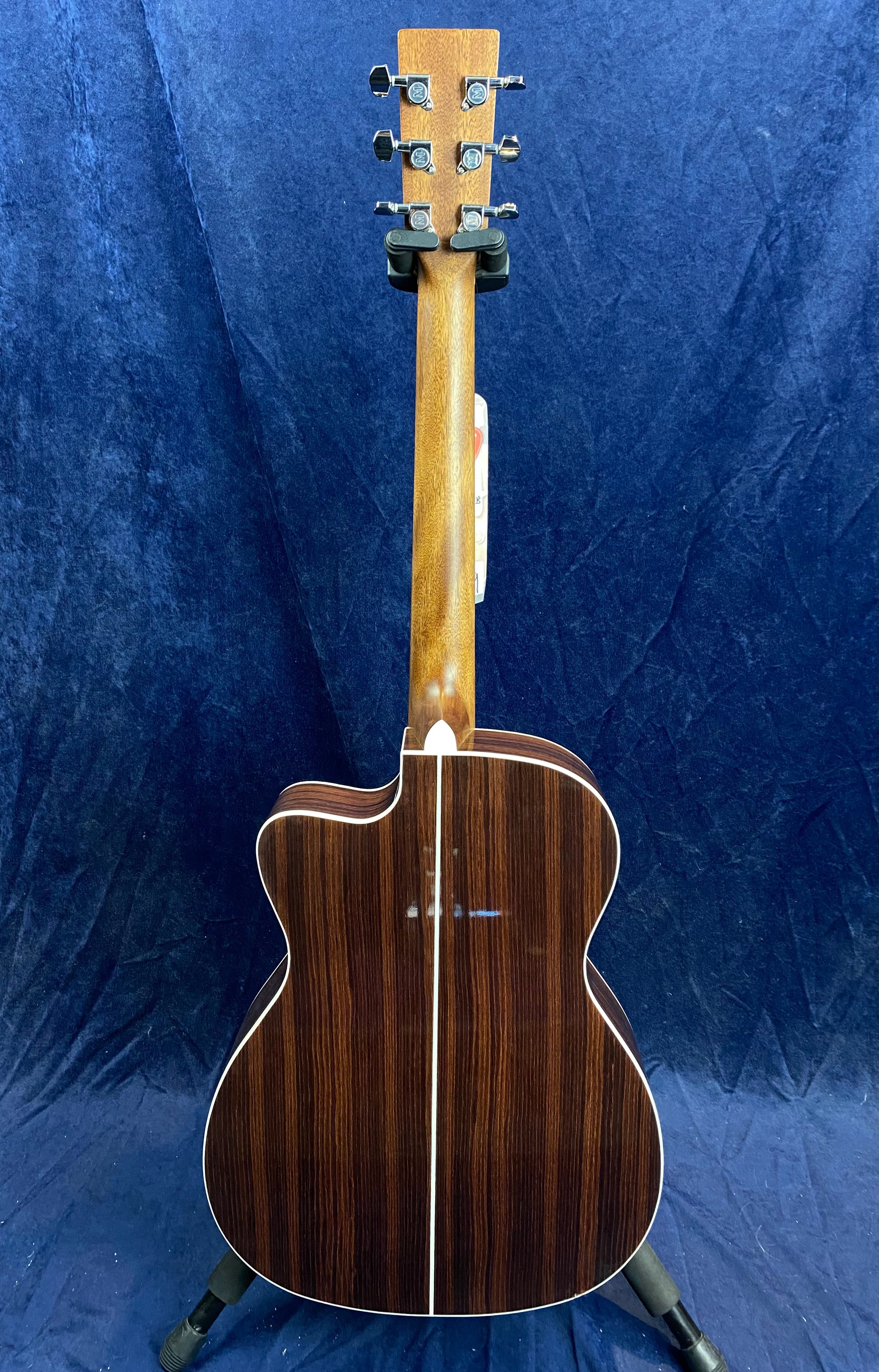 Acoustic guitar viewed from the back, showing a dark wood body with a pale center stripe, light wood neck, and six chrome tuning pegs.