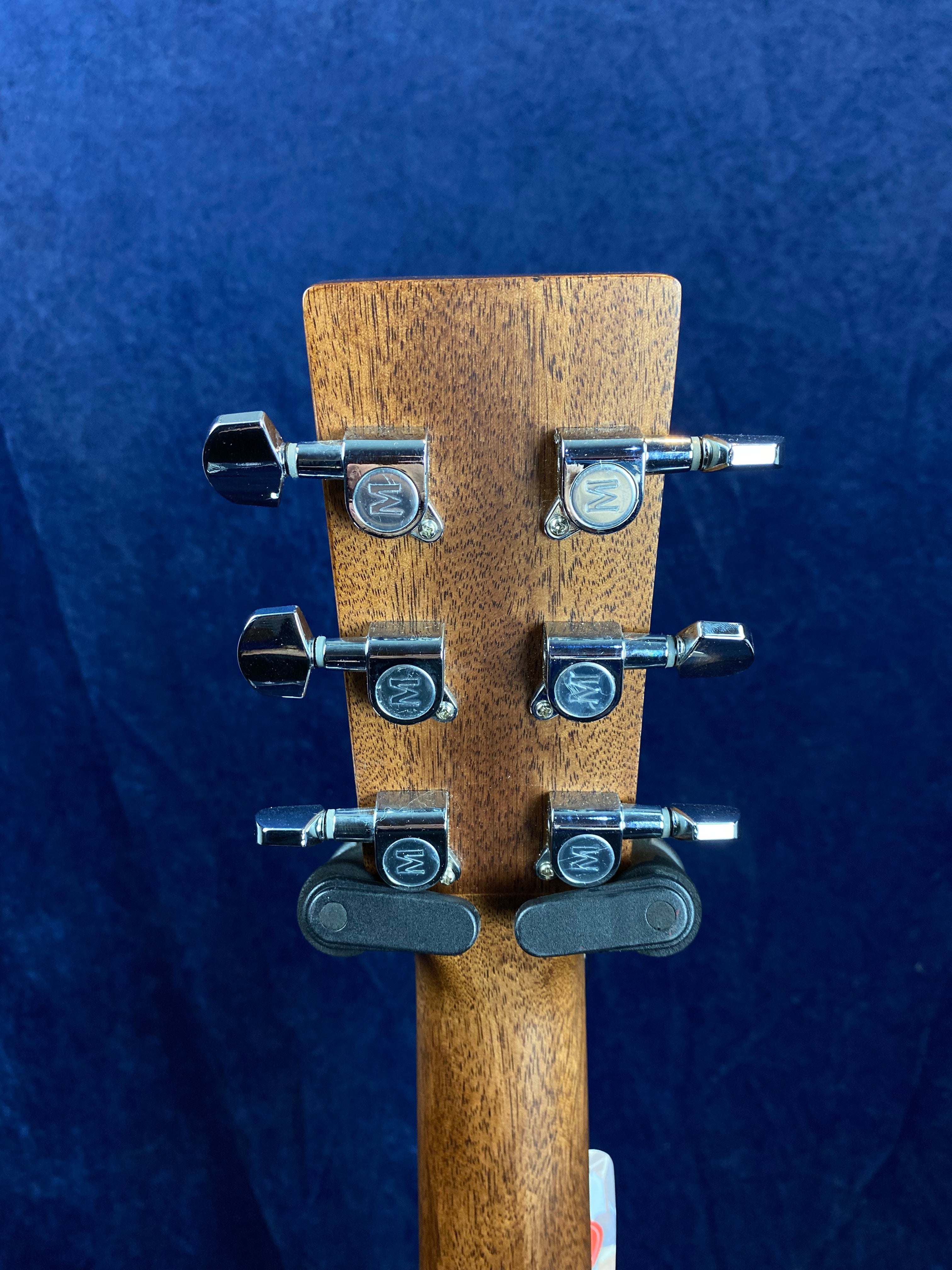 Six chrome tuning pegs with "M" logos on the wooden headstock of an acoustic guitar, two capos clipped to the back of the headstock.