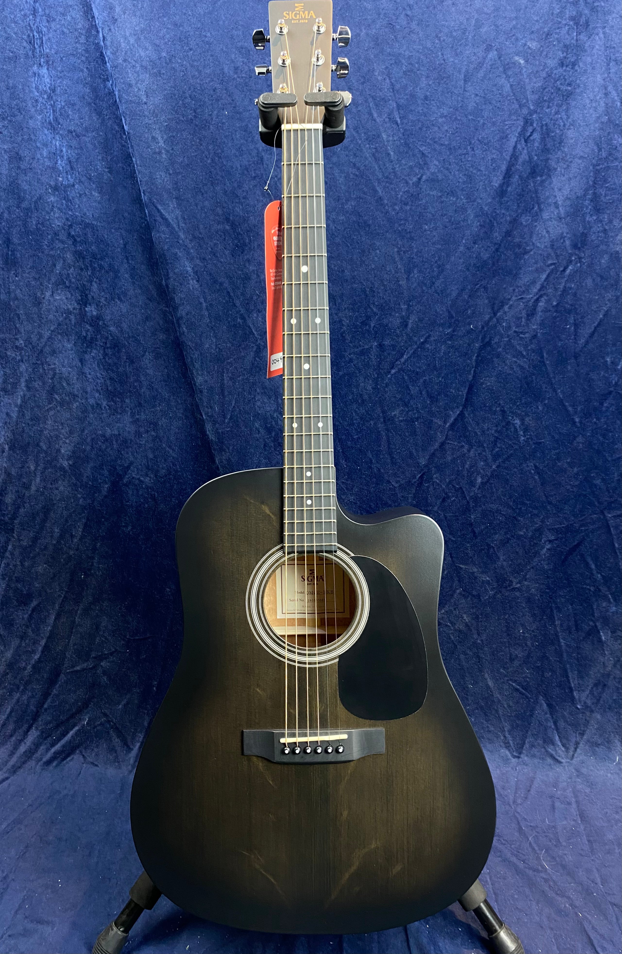 Black acoustic guitar with a matte finish, white dot inlays on the fretboard, a round sound hole, and a black pickguard, displayed vertically on a stand.