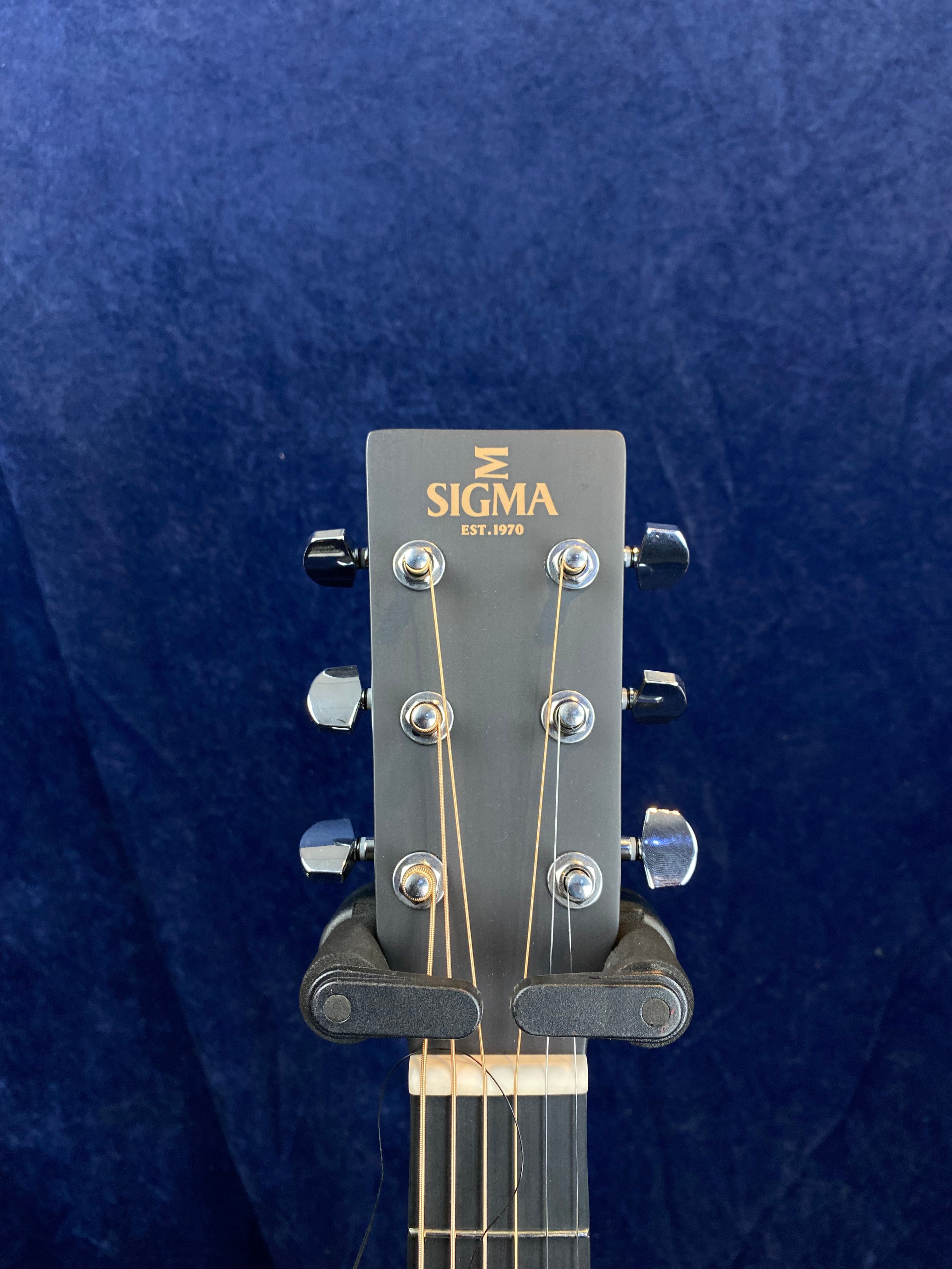 Headstock of a Sigma acoustic guitar with six tuning pegs, gold logo, and strings, held upright in a black guitar stand against a dark blue background.