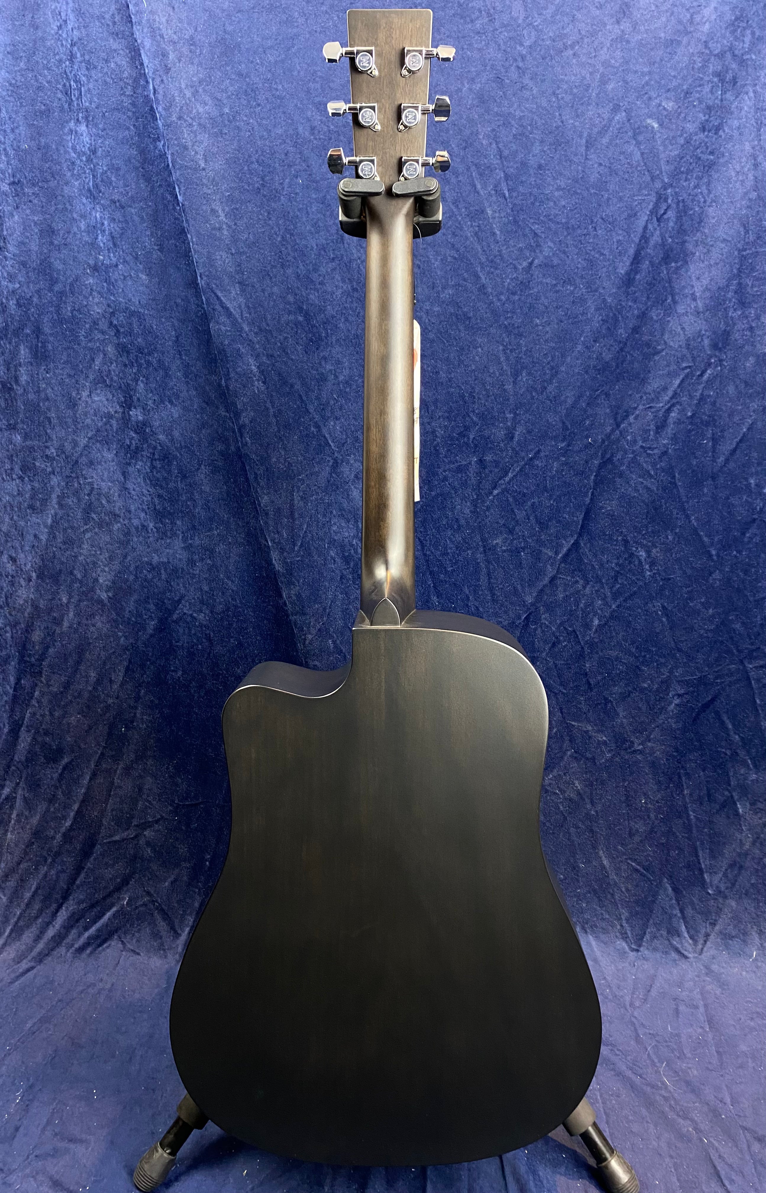 Back view of a matte dark wood acoustic guitar with silver tuning pegs, cutaway body, and a smooth neck against a dark blue fabric background.