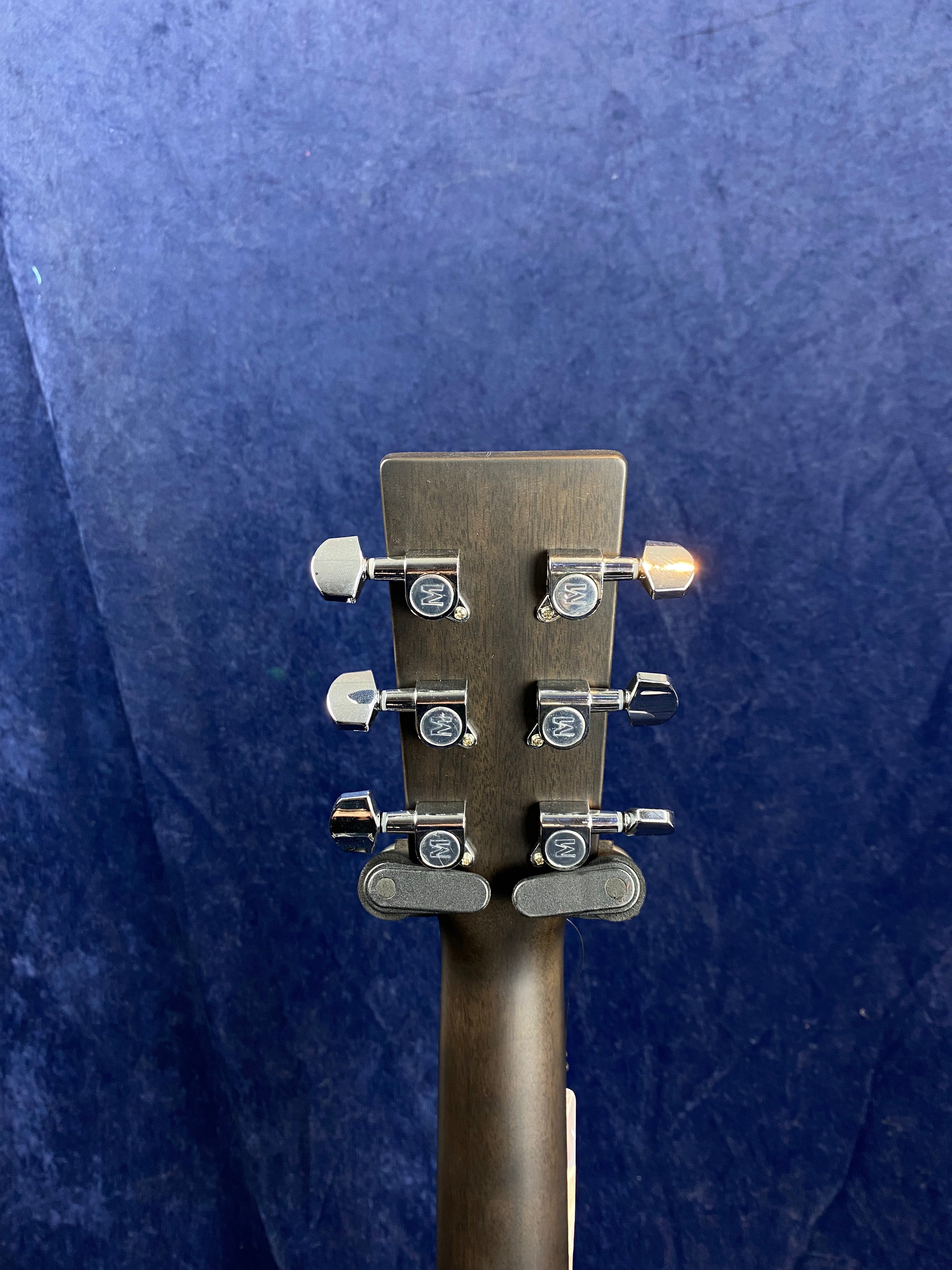 Back of an acoustic guitar headstock showing six chrome tuning pegs and a dark wood finish, set against a dark blue background.