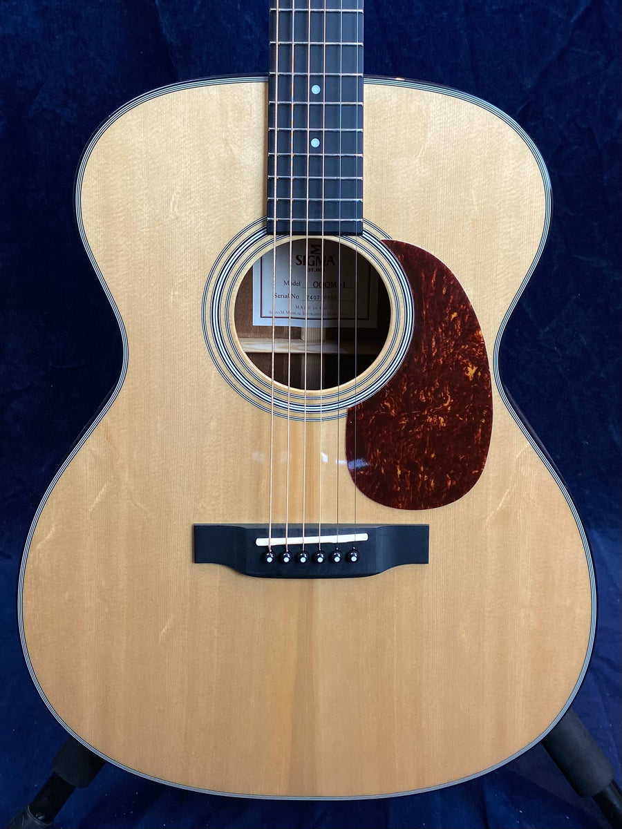 Light wood acoustic guitar with six strings, a black bridge, round sound hole, and a brown tortoiseshell-style pickguard on the lower right side.