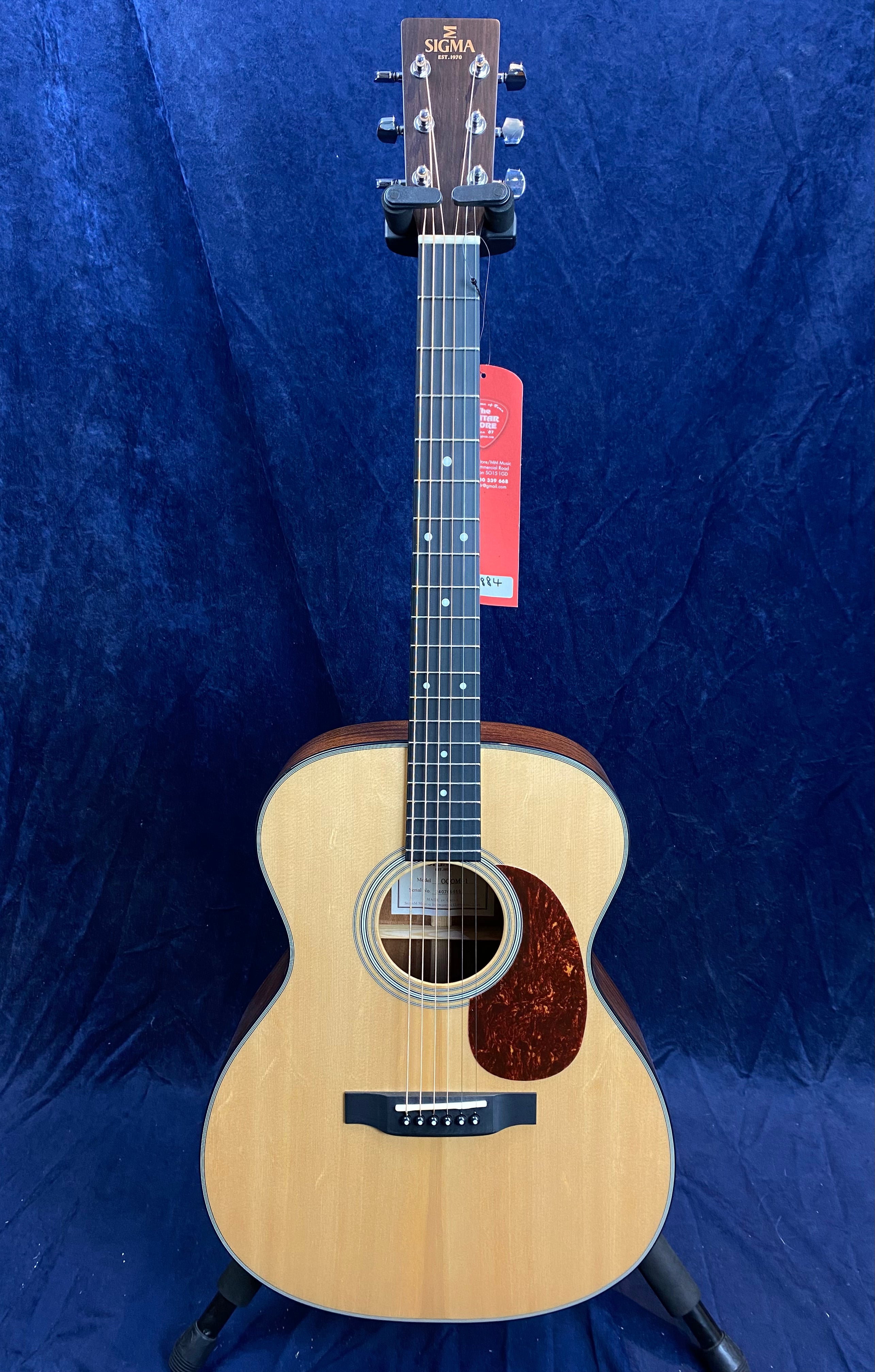 Sigma acoustic guitar with a natural wood finish, dark pickguard, six strings, and a label hanging from the tuning pegs.
