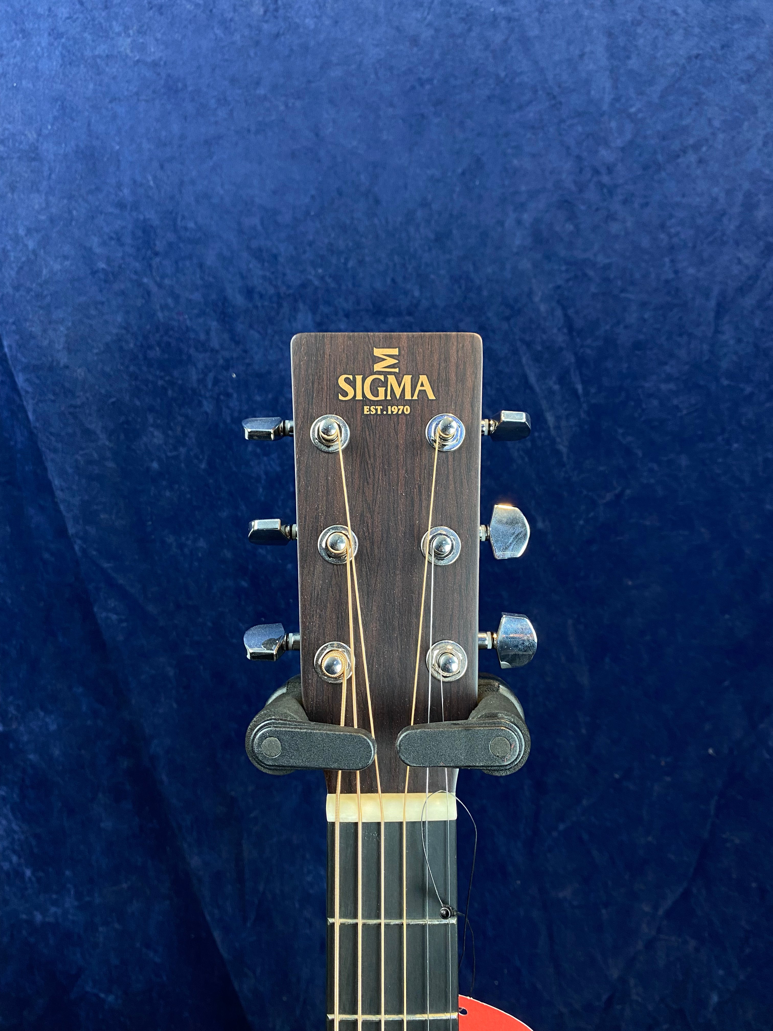 Sigma acoustic guitar headstock with six tuning pegs, logo and "Est. 1970" text, held by a black guitar hanger against a dark blue background.