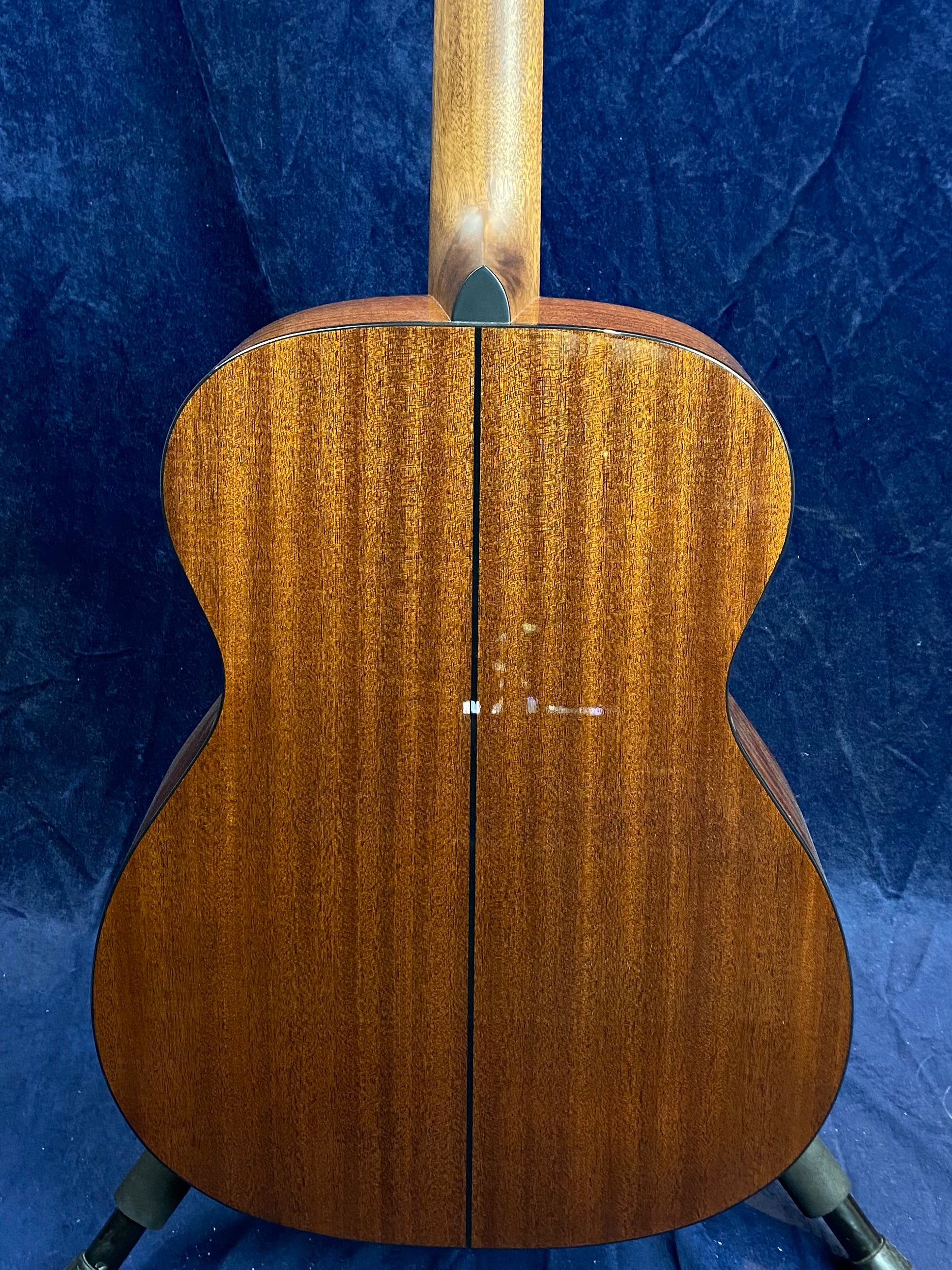 Mahogany acoustic guitar back with a straight center seam and natural wood grain, resting on a stand against a dark blue background.