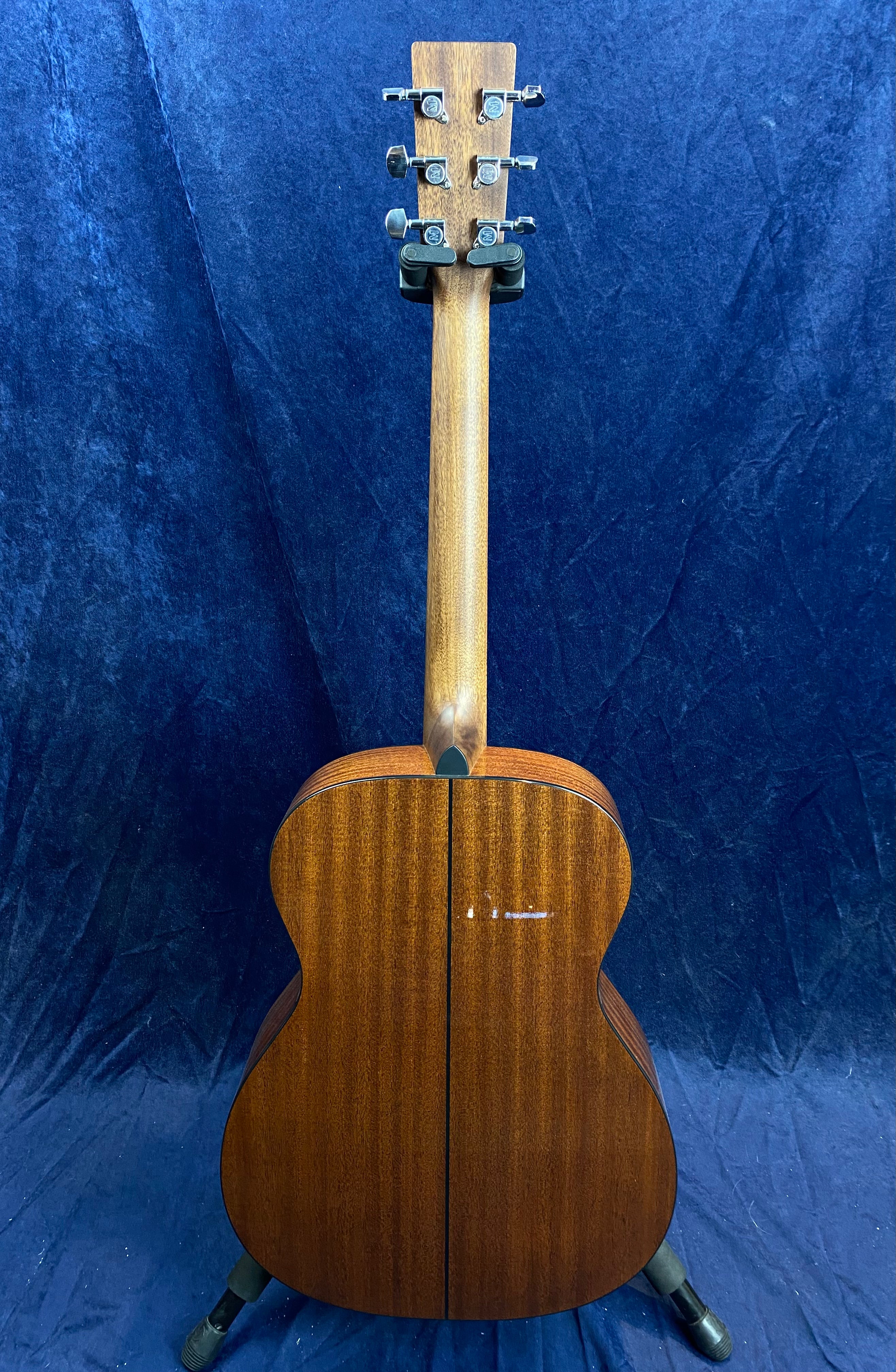 Brown acoustic guitar shown from the back, featuring a wooden finish with a visible center seam and silver tuning pegs on the headstock.