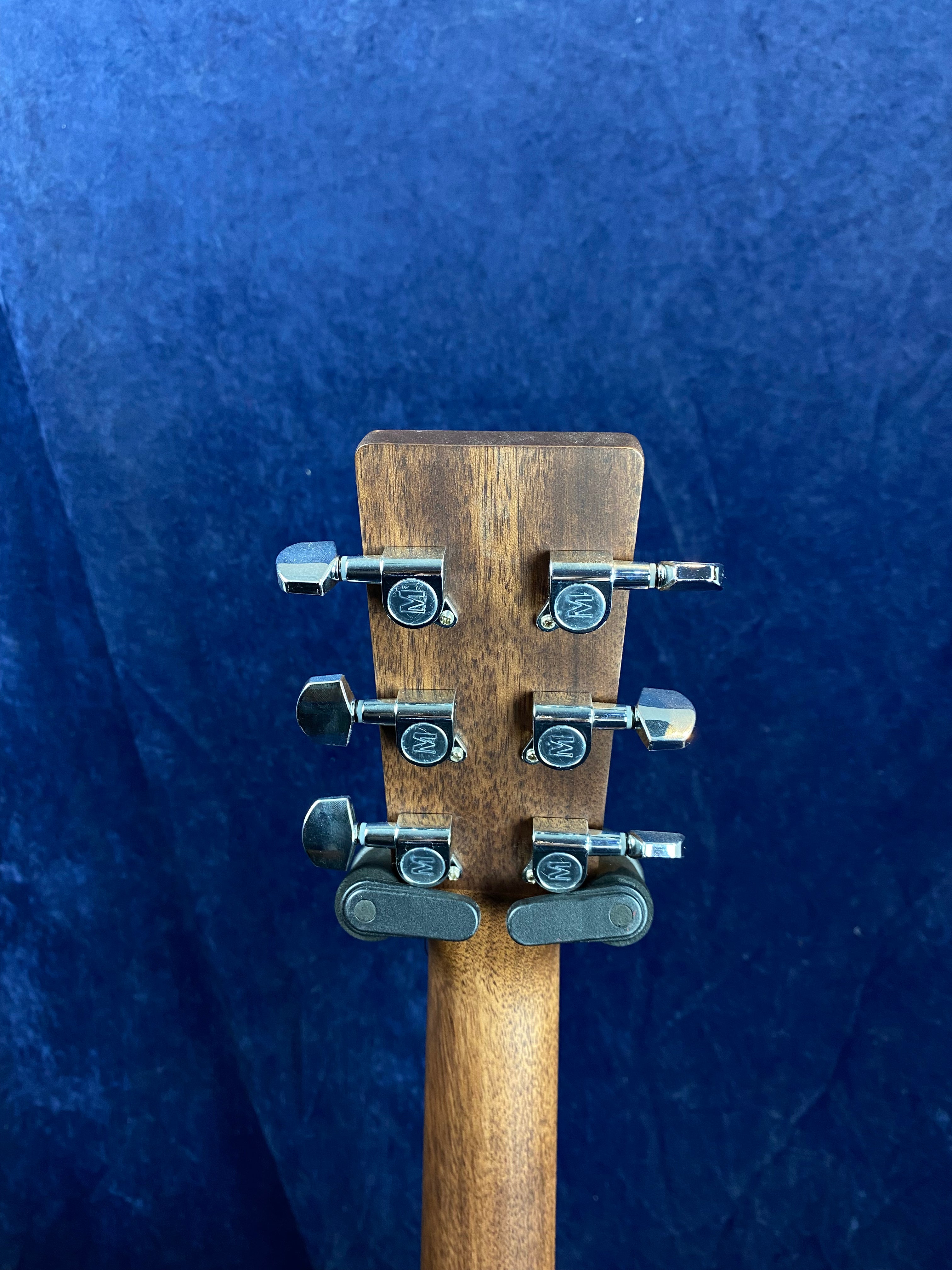 The back of an acoustic guitar headstock with six silver tuning pegs against a dark blue background.