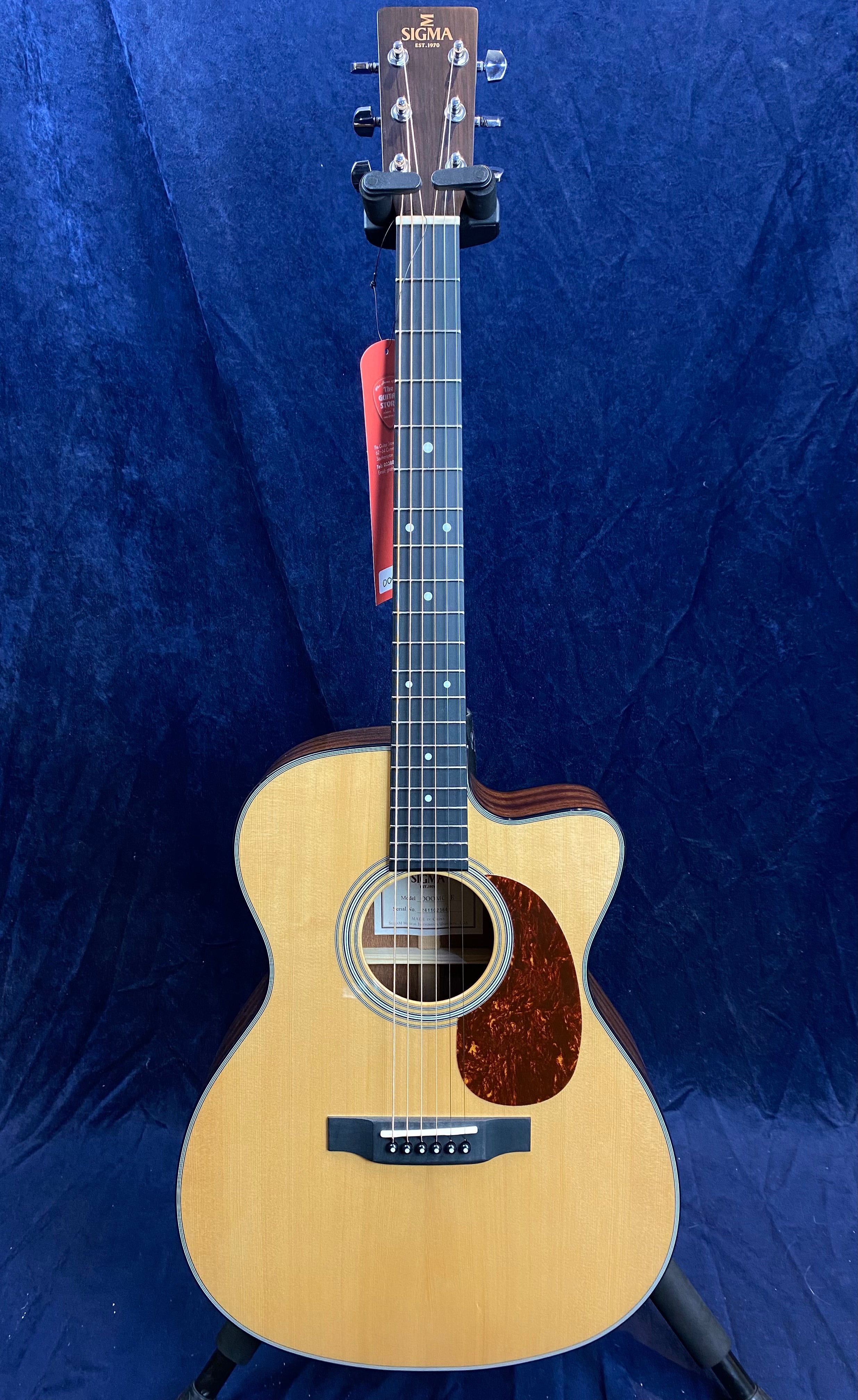 Sigma acoustic guitar with a natural wood finish, dark pickguard, cutaway body, six strings, and dot inlays on a dark fretboard, standing upright.