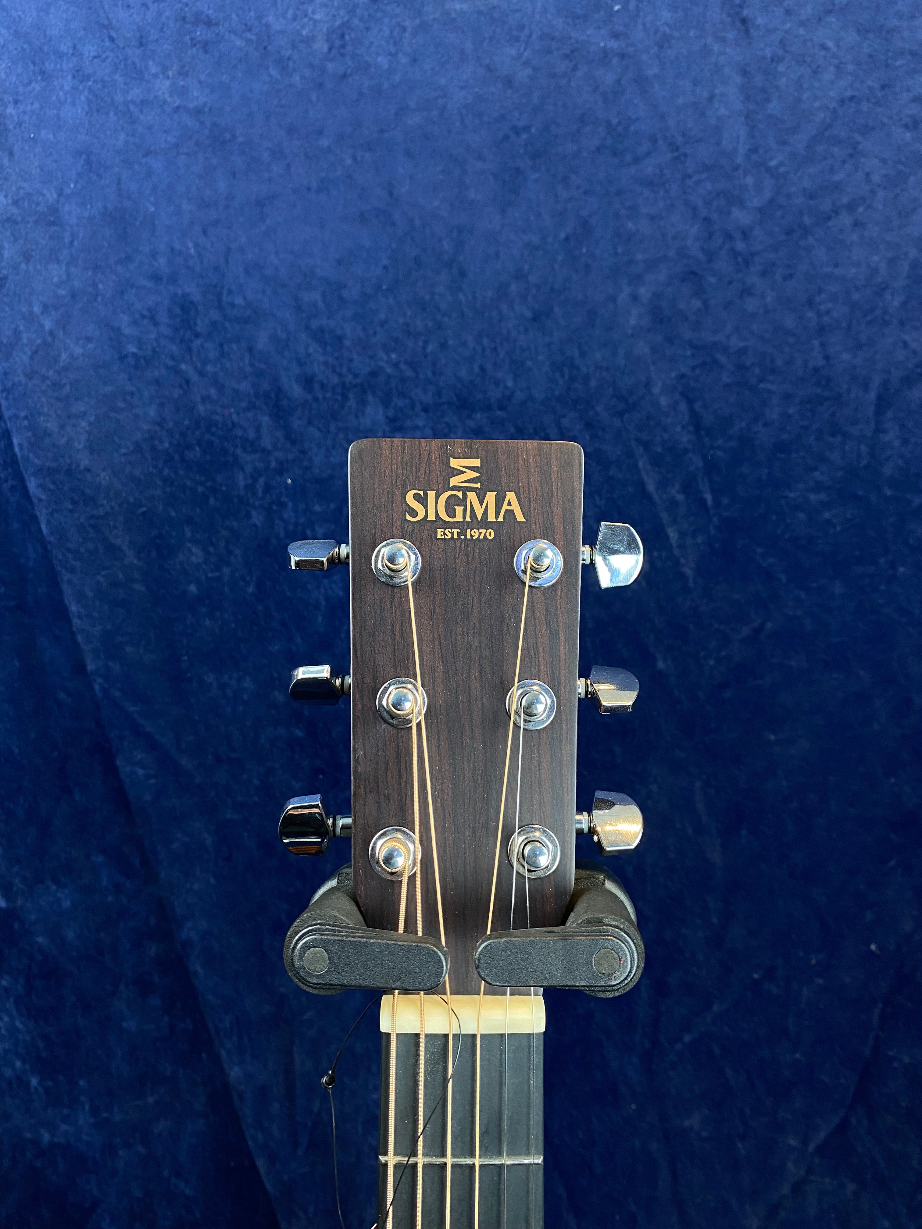Headstock of a Sigma acoustic guitar with six tuning pegs, wood grain finish, and gold “SIGMA EST. 1970” logo against a dark blue background.