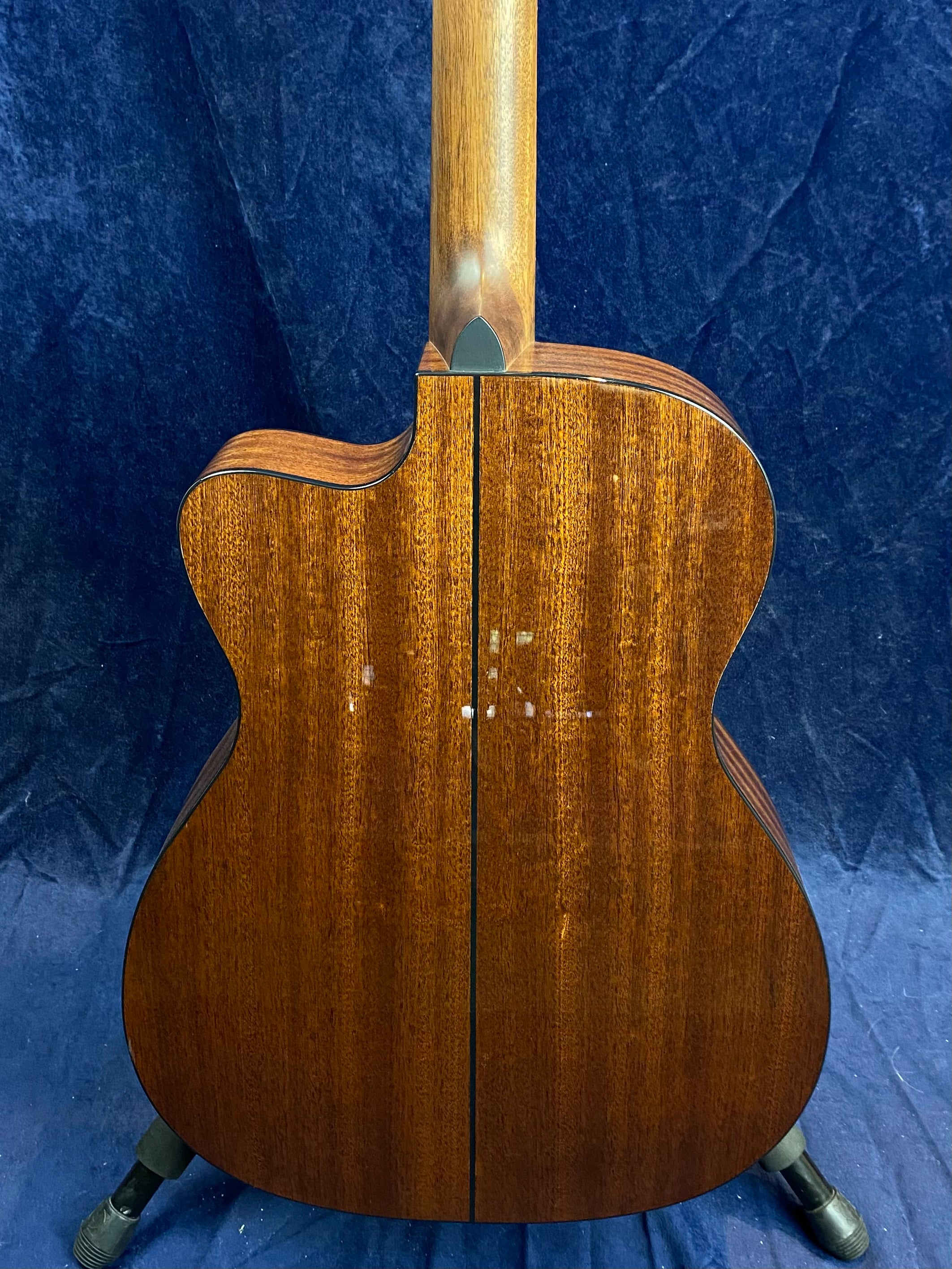 Back view of an acoustic guitar with a rich brown wood finish, a center seam, and a smooth neck transition, standing on a black tripod stand.