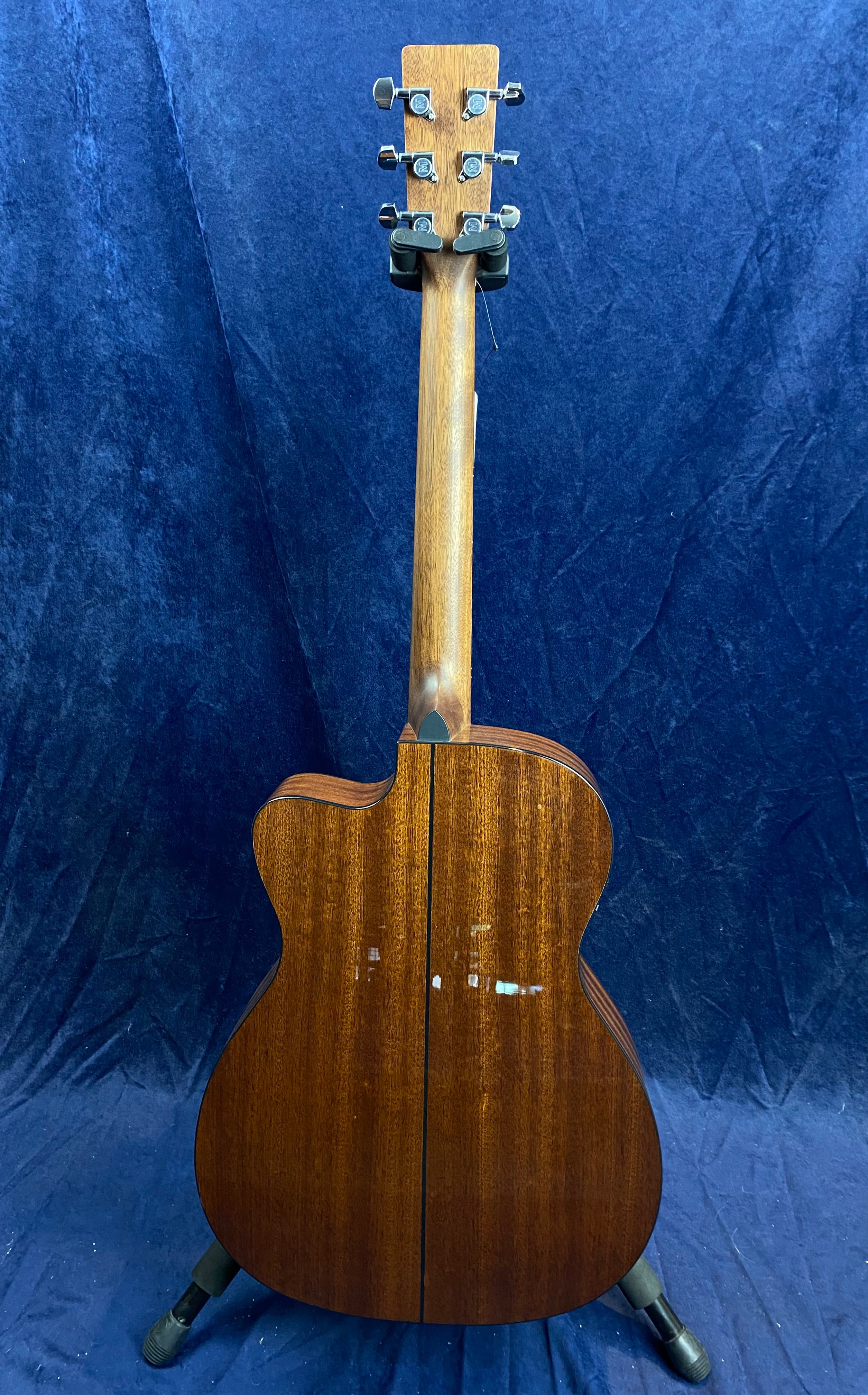Mahogany acoustic guitar shown from the back, featuring a cutaway body and chrome tuning pegs, standing on a floor stand against a blue backdrop.