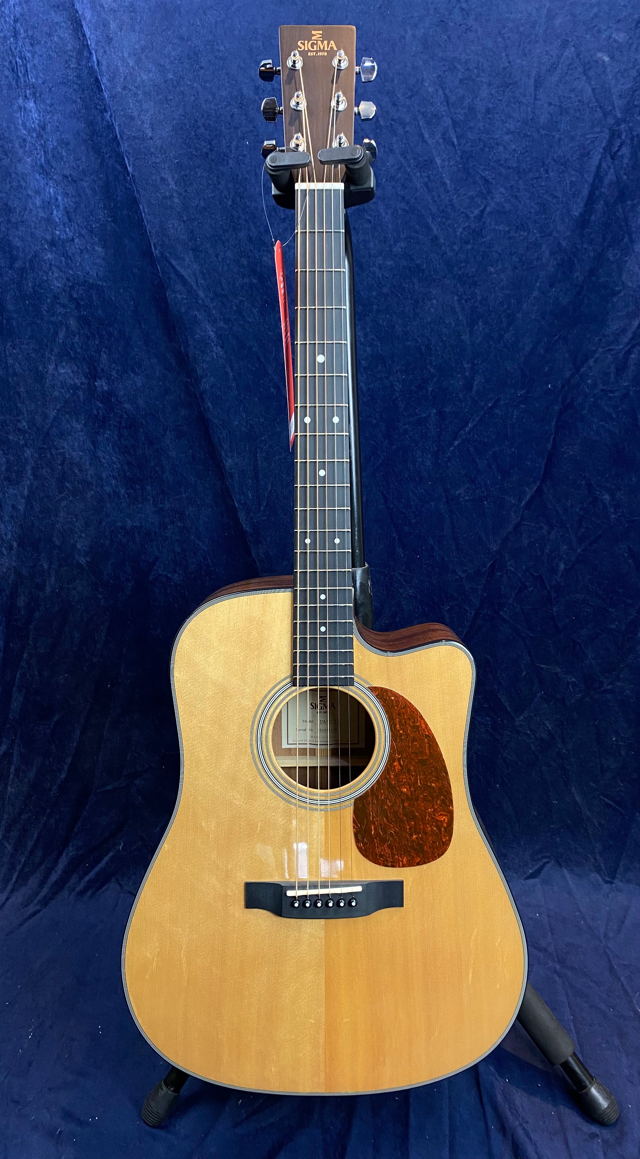 Sigma acoustic guitar with a natural wood finish, dark pickguard, cutaway design, and six strings, standing on a black tripod stand.