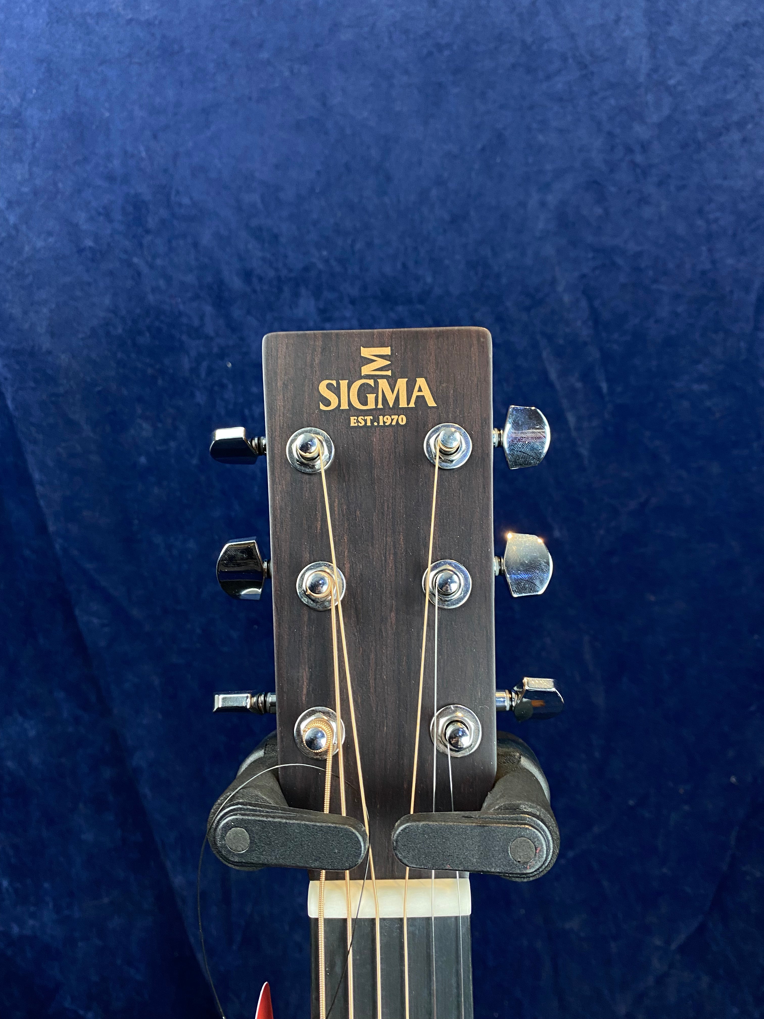 The headstock of a Sigma acoustic guitar shows six tuning pegs, with the Sigma logo and "EST. 1970" printed in gold lettering.