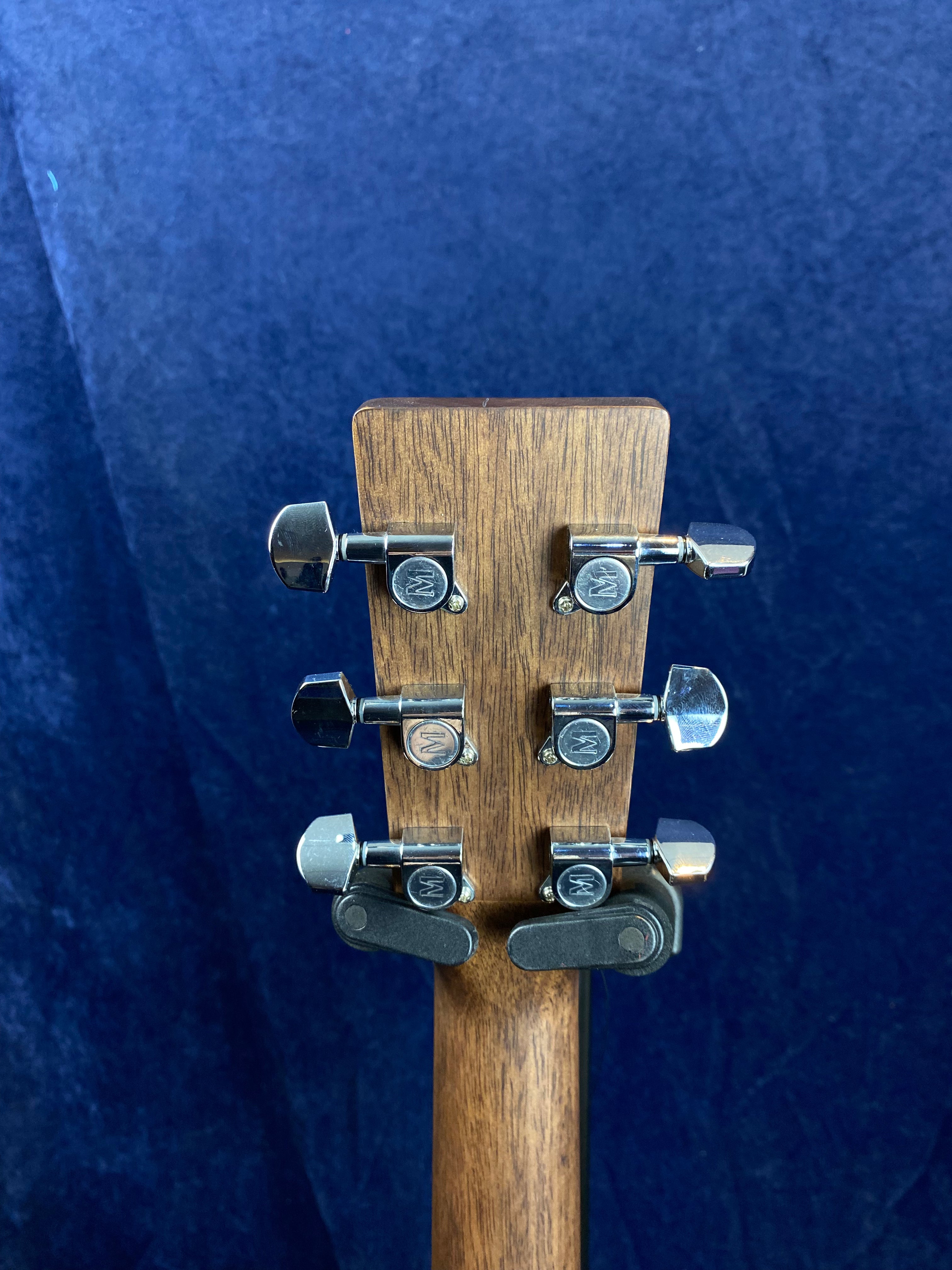 Back of an acoustic guitar’s headstock showing six chrome tuning pegs on a natural wood finish, against a dark blue background.