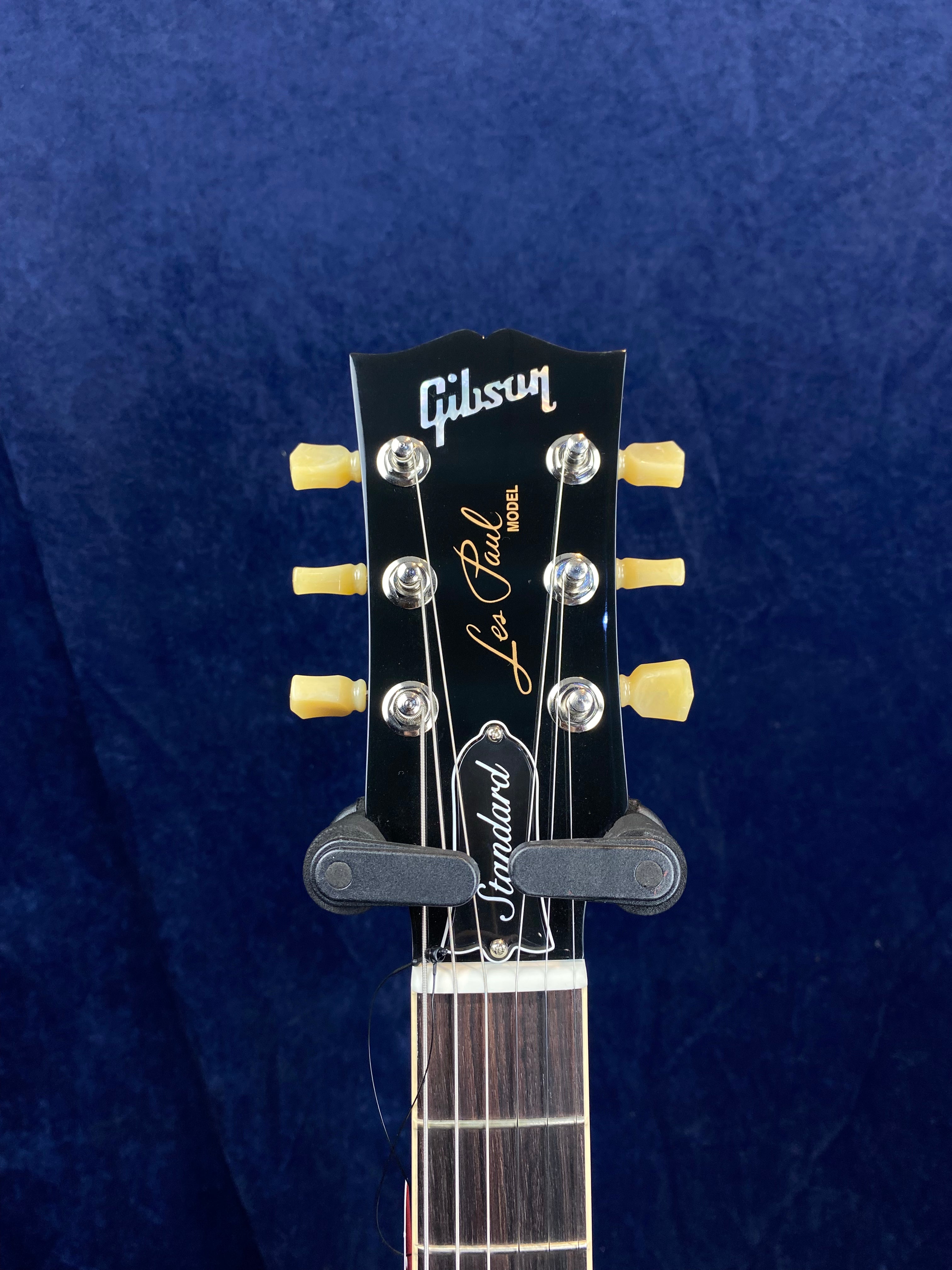 Close-up of a Gibson Les Paul electric guitar headstock with cream tuning pegs and "Standard" truss rod cover against a dark blue background.